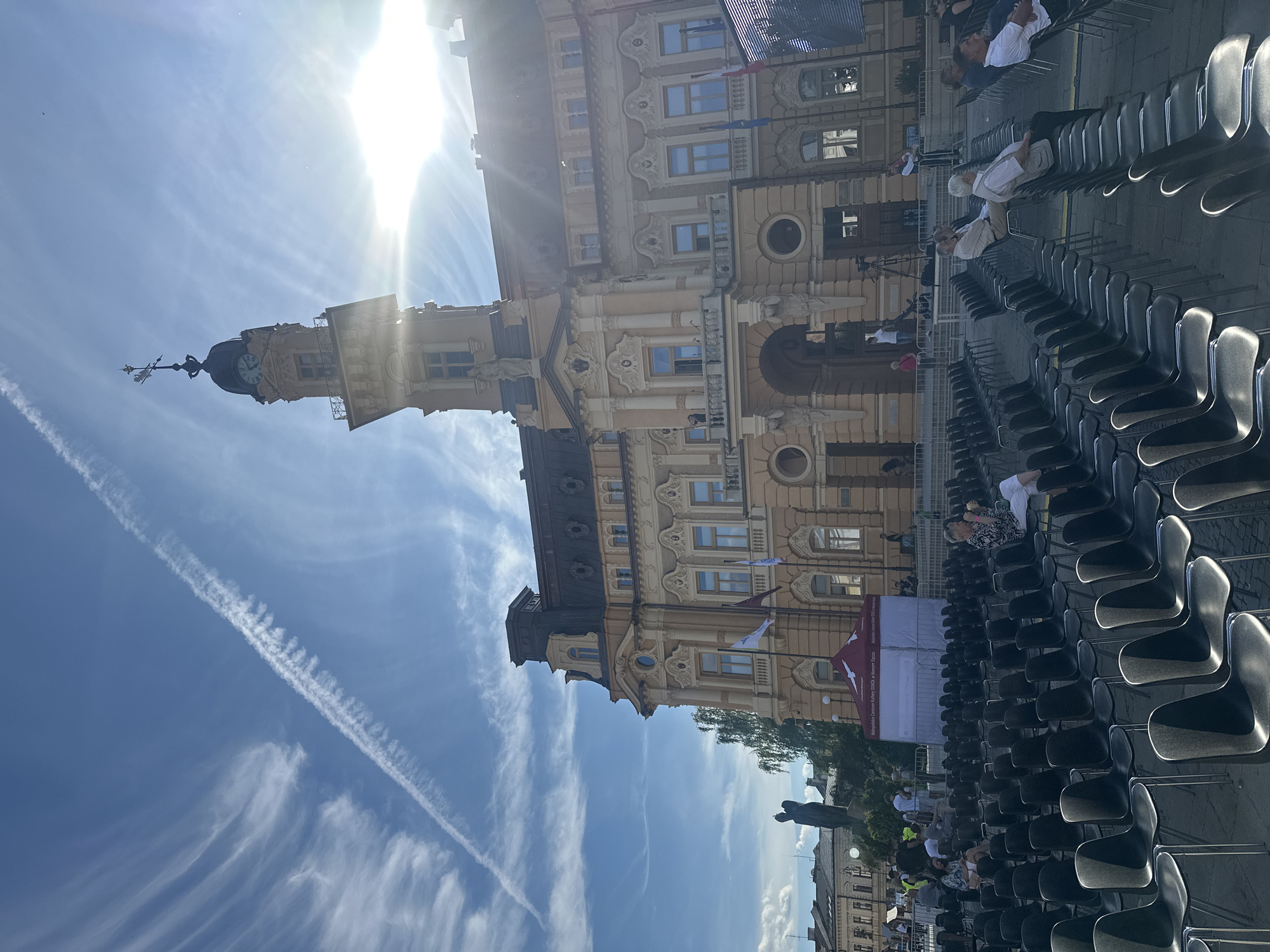 The image shows an outdoor event setup in front of the Sokół building in Nowy Sącz, Poland. Rows of empty black chairs fill the foreground, with some people seated in them in the middle ground. The Sokół building, an ornate yellow structure with a clock tower, dominates the background. The sky is blue with wispy clouds and a prominent contrail. A red and white tent is set up in front of the building, and a statue of a figure is visible to the left of the building. The sun is shining brightly, creating lens flare in the upper right portion of the image.