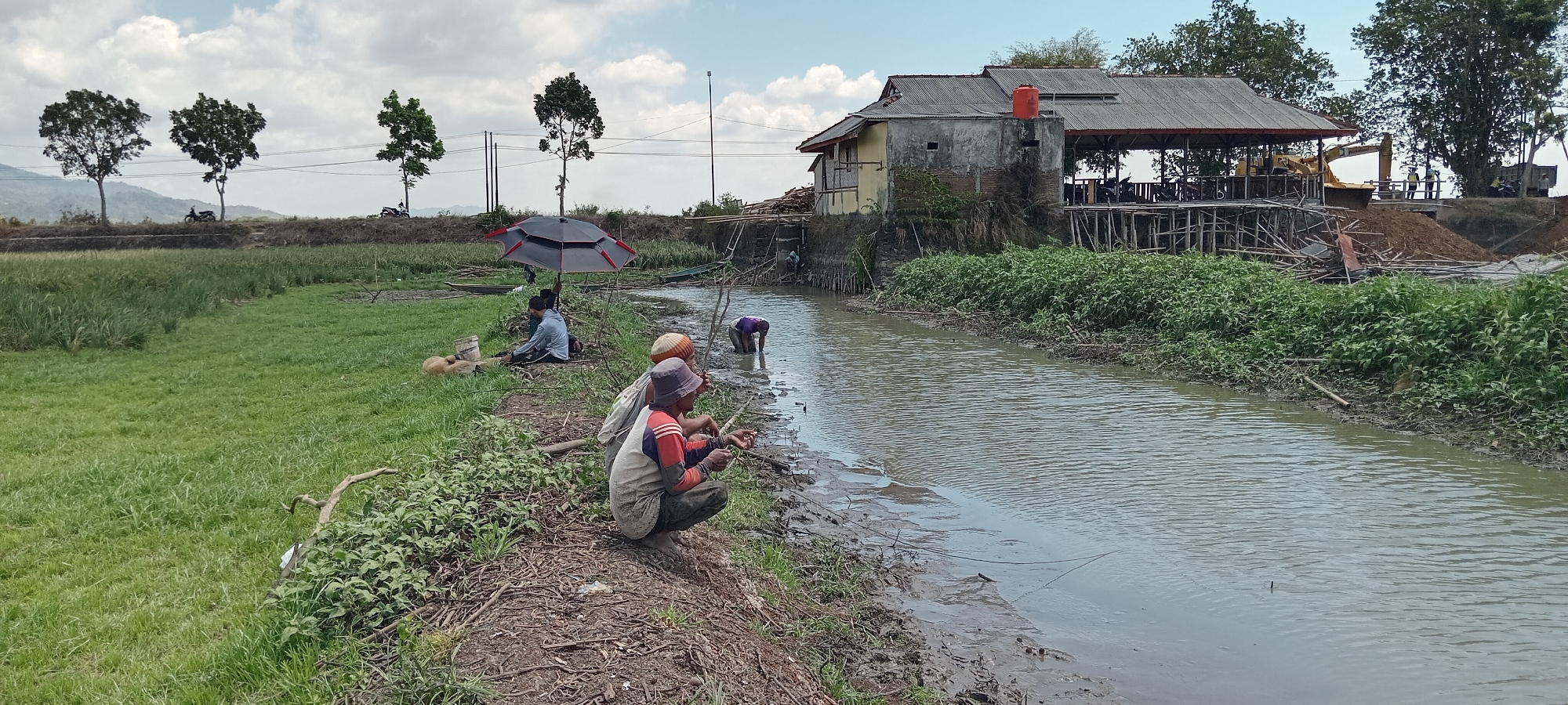 In Paledah, Indonesia, a muddy canal flows through a rural landscape. On the left bank, lush green grass meets the muddy bank, where several individuals are engaged in fishing. One person, wearing a blue shirt and white pants, sits under a black and red umbrella, holding a fishing rod. To their right, another person in a grey and red shirt crouches, also fishing. A third person in a purple shirt is partially submerged in the canal, appearing to be tending to something in the water. Further up the bank, near a dilapidated building with a red water tank on its roof, a yellow excavator is visible, suggesting ongoing construction or land alteration. Behind the building, wooden scaffolding and piles of dirt indicate a work site. The background features a line of trees, distant hills, and a cloudy sky.