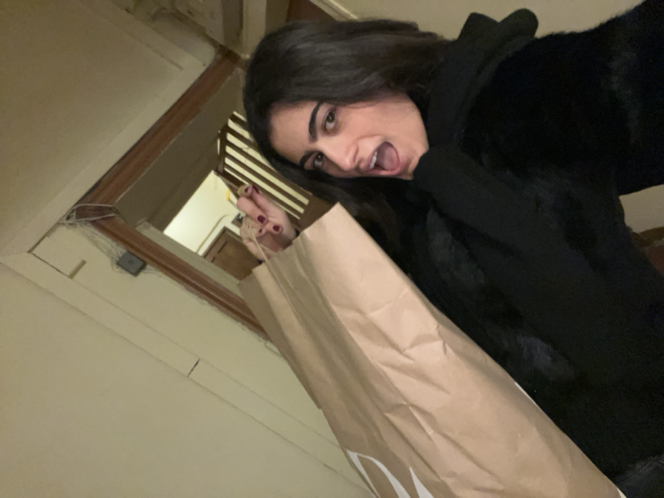 A woman with dark hair and a black fuzzy jacket poses with her mouth open wide, holding a brown shopping bag. Her manicure is visible. She looks at the camera at an angle. The background features a white wall and doorframe with exposed wiring. The view into the room behind her includes light-colored wooden furniture and white vertical slats. The photo seems to be taken inside a house or apartment.