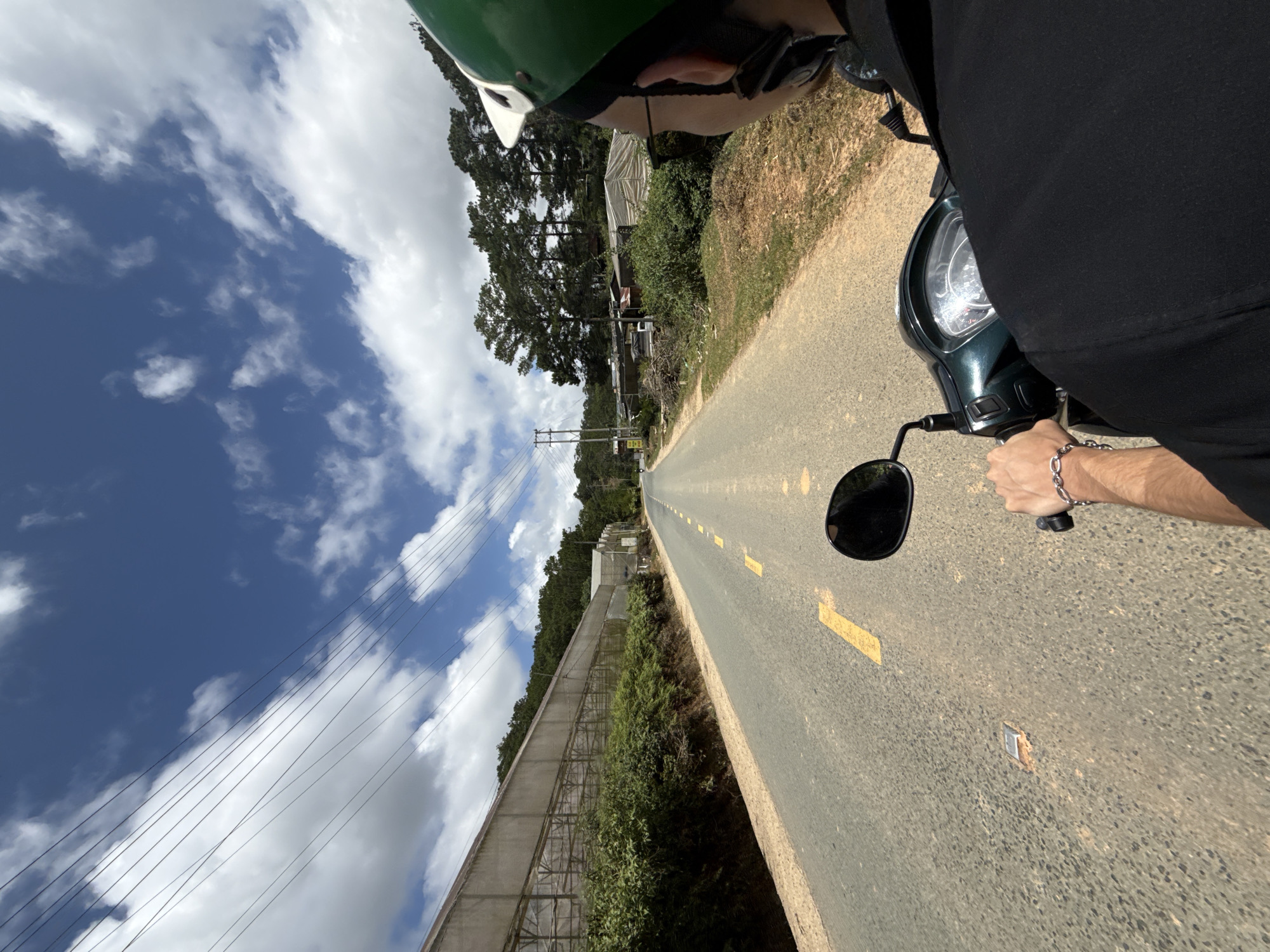 A first-person perspective shot shows a person riding a dark green scooter down a paved road in Lạc Dương, Vietnam. The road is lined with yellow painted markings and is flanked by lush greenery on the left and pine trees on the right. Large agricultural greenhouses with translucent coverings are visible on the left side of the road, extending into the distance. The sky is a vibrant blue with scattered white clouds and power lines crisscrossing overhead. The rider is wearing a green helmet and a black shirt, with a silver bracelet on their left wrist. A silver handlebar is visible, and a side mirror on the scooter reflects the road ahead. The background features a cluster of buildings and trees.