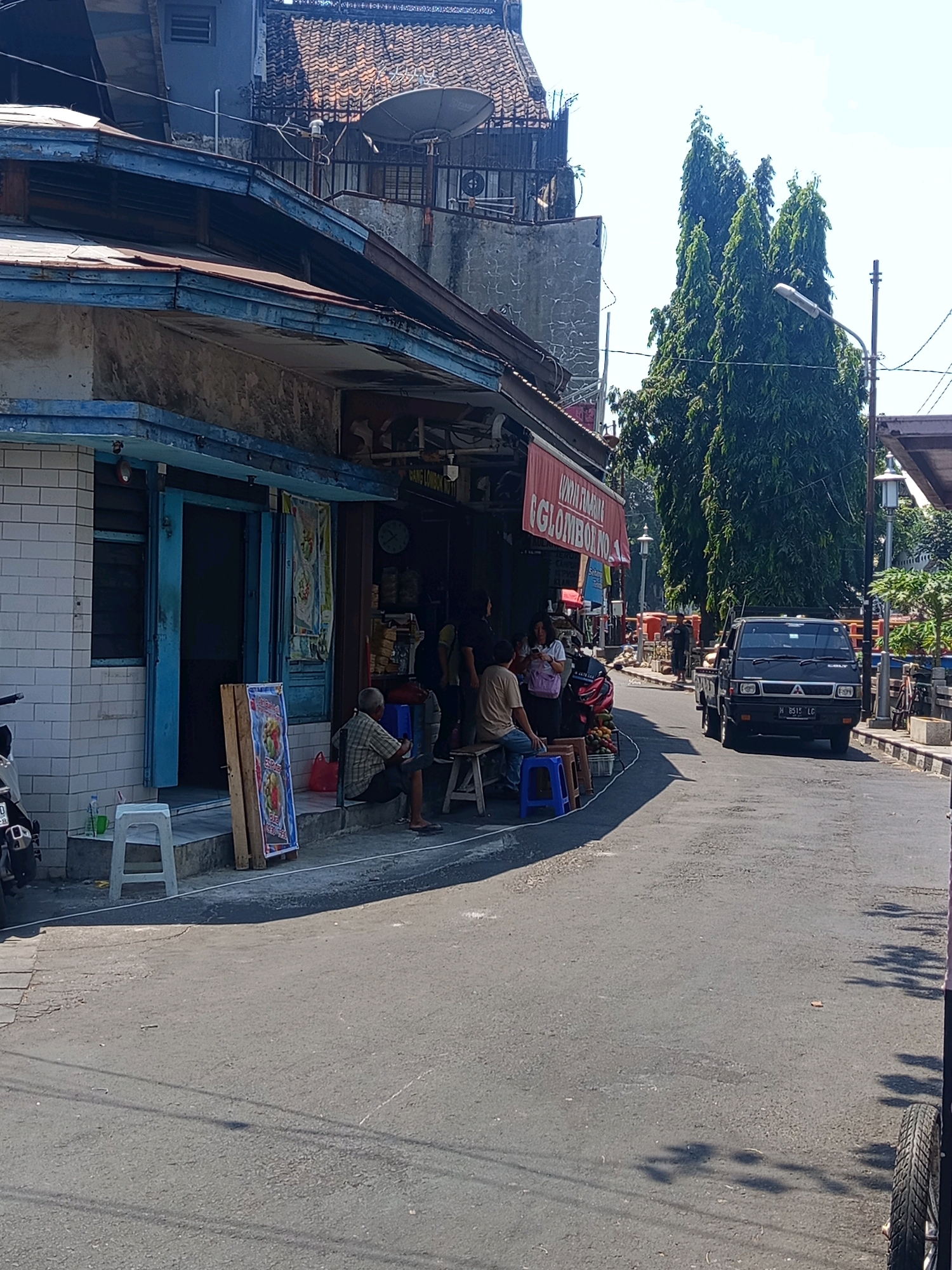 The image depicts a street scene in Semarang, Indonesia, likely a local market or commercial area. The foreground is an asphalt road with a few shadows cast across it. On the left side of the road, a weathered building with a blue awning and white tiled walls houses a shop. A blue door stands open, and a person is seated on a stool outside. A colorful poster is propped against the wall. Further down the street, several people are gathered near another shop entrance. A red awning with text is visible, and a clock hangs on the wall. To the right of the street, a black pickup truck is parked, with its license plate clearly visible. Tall, slender trees line the right side of the road, and a street lamp is present. The overall impression is of a bustling, ordinary day in a city environment.