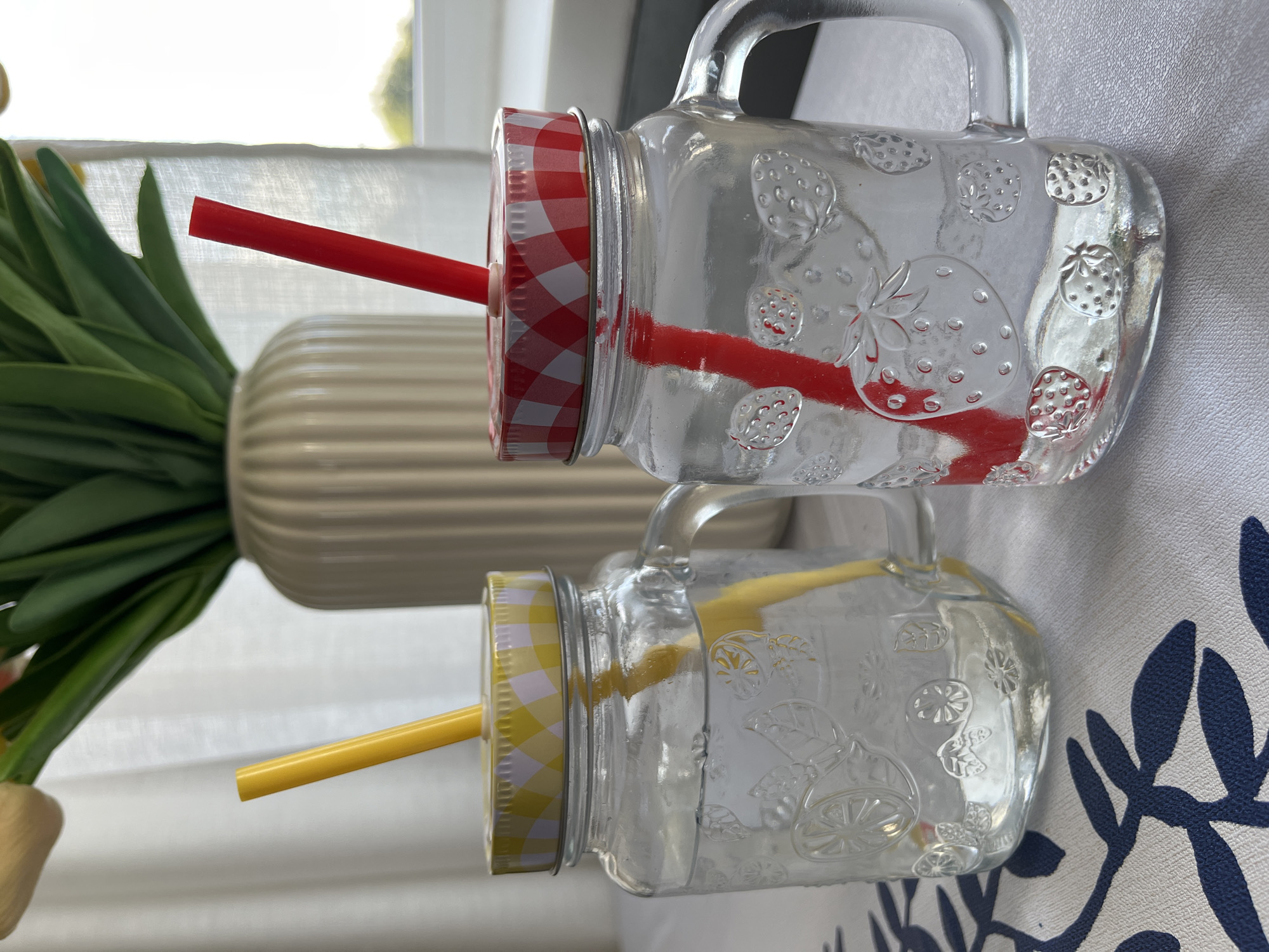 Two clear glass mason jar mugs with handles are placed on a white tablecloth with a blue floral pattern. Each mug is filled with a clear liquid and has a metal lid with a straw. The mug on the left has a yellow and white checkered lid and a yellow straw. The mug on the right has a red and white checkered lid and a red straw. The glass of both mugs is embossed with fruit designs, the left with citrus slices and leaves, and the right with strawberries. In the background, a light-colored vase with green foliage and yellow flowers is visible. The scene is illuminated by natural light, suggesting it is daytime. There are no people present in the image. The location of this scene is not identifiable from the image itself and cannot be confirmed to be Kohtla-Järve linn, Estonia.