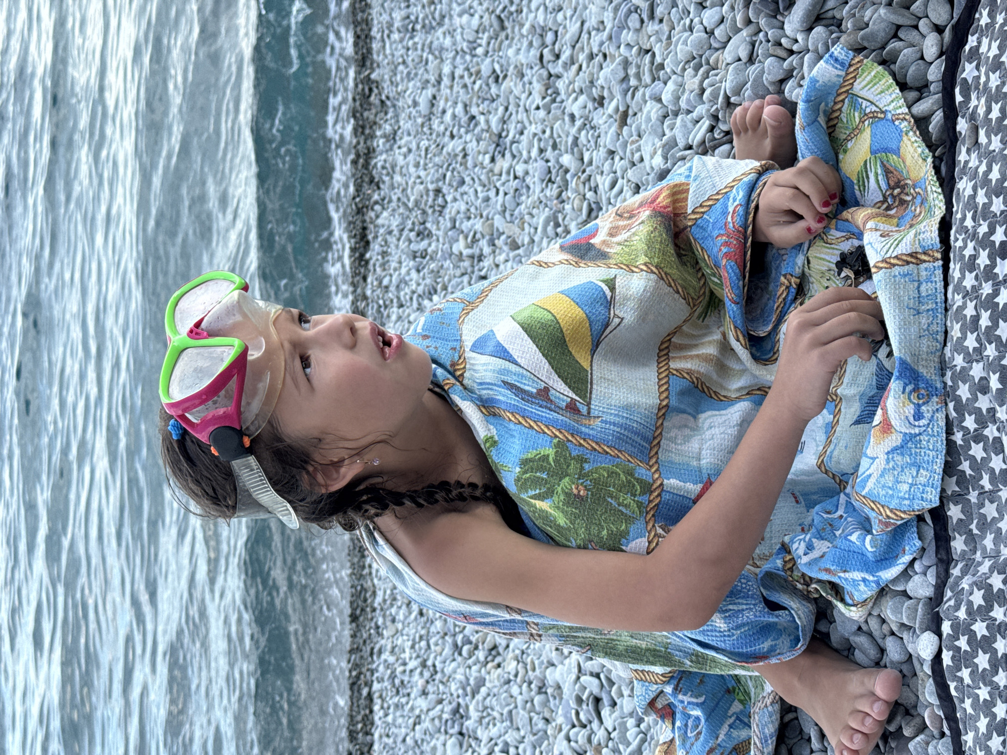 A young girl is lying on a pebble beach, wrapped in a towel with a nautical print. She is wearing a snorkeling mask with bright green frames and a pink strap, which is pushed up onto her forehead. The towel features images of sailboats, palm trees, and fish. Her feet are visible at the bottom of the frame, resting on the towel. To her left, the ocean stretches out, with gentle waves lapping against the shore. The sky appears overcast, suggesting it might be a cloudy day. The scene conveys a sense of relaxation and readiness for water activities.
