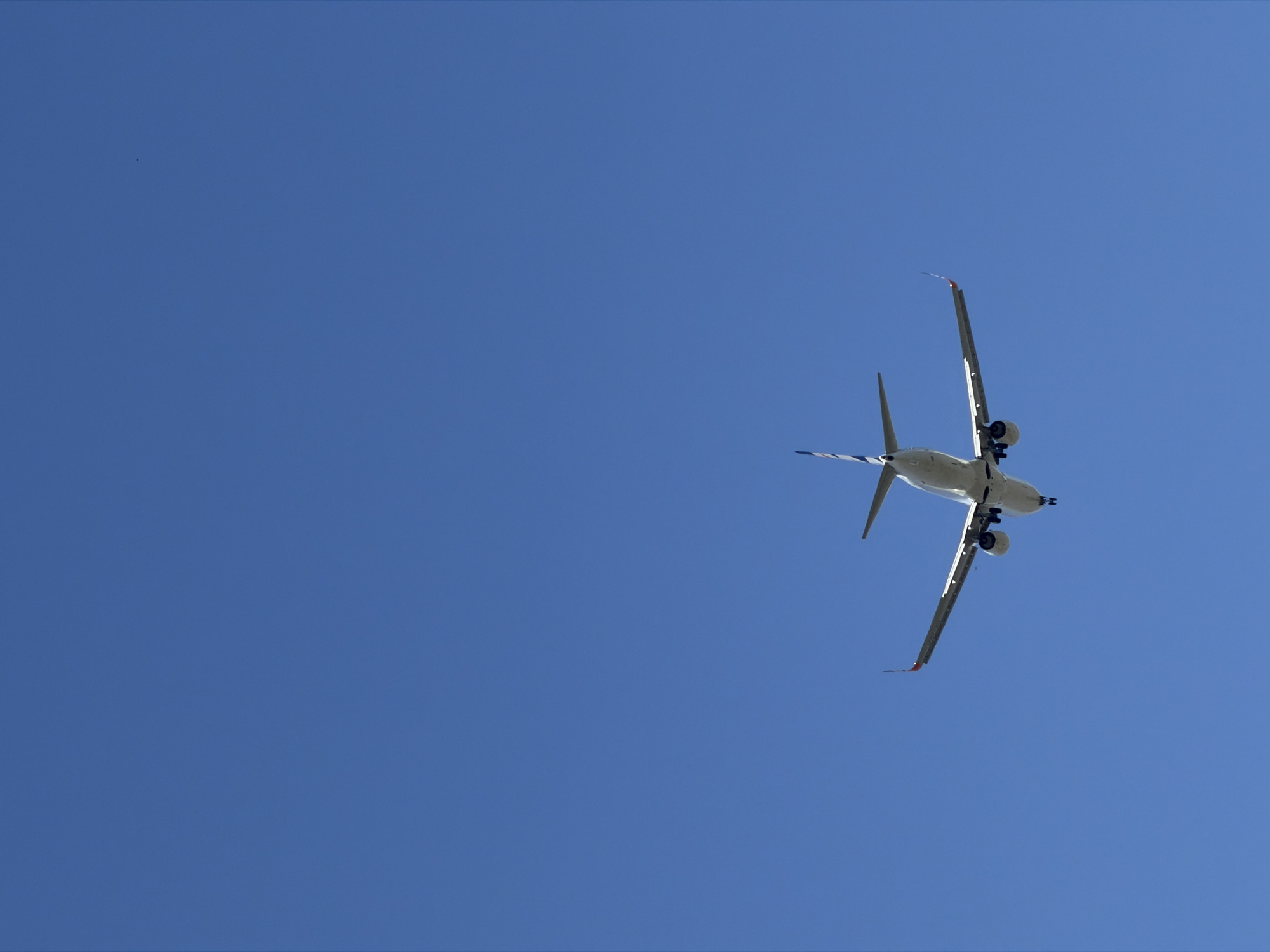 This media file captures an airplane in flight against a clear, bright blue sky. The perspective is from below, looking up at the aircraft as it soars overhead. The airplane is predominantly white with some blue and red accents visible on its tail fins and wingtips. Its landing gear appears to be down, suggesting it might be preparing to land or has just taken off. The sun is high in the sky, casting minimal shadows and indicating it is likely midday. The overall setting is an open sky, likely over Burgas, Bulgaria, given the location context. There are no people or discernible structures in the frame, emphasizing the vastness of the sky and the solitary presence of the airplane. No text is visible on the aircraft or in the environment.