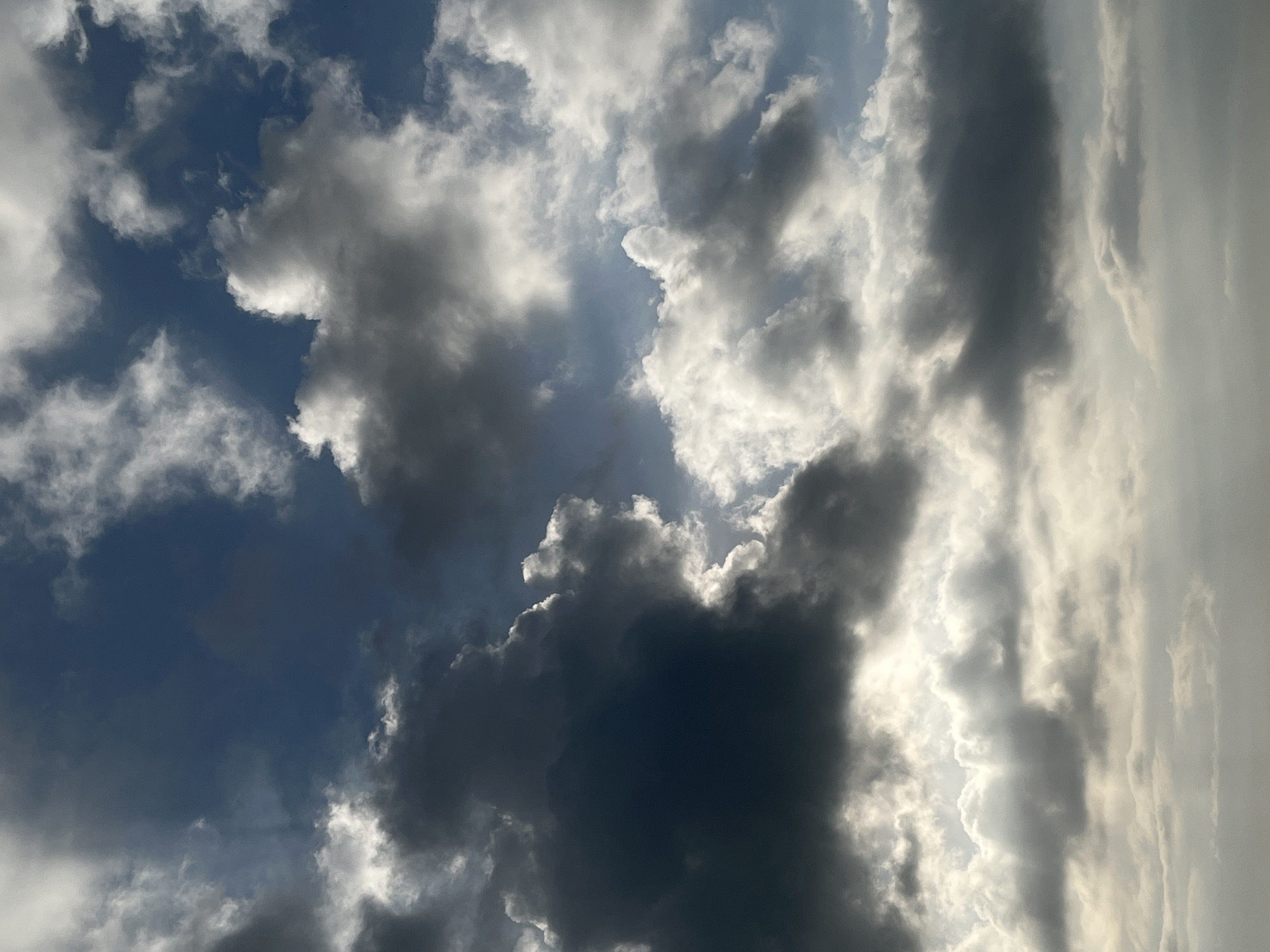 The image is a low-angle shot of a cloudy sky with the sun peeking through. The clouds range in color from dark grey to bright white, creating a dramatic contrast. Some clouds appear wispy and translucent, while others are dense and voluminous. The sunlight filters through in rays, illuminating portions of the clouds and casting a soft glow on the sky. There are no people, subjects, objects, or actions depicted in this image; it is solely a view of the sky and clouds. The scene does not provide any specific indicators of the city of Tambakreja, Indonesia.