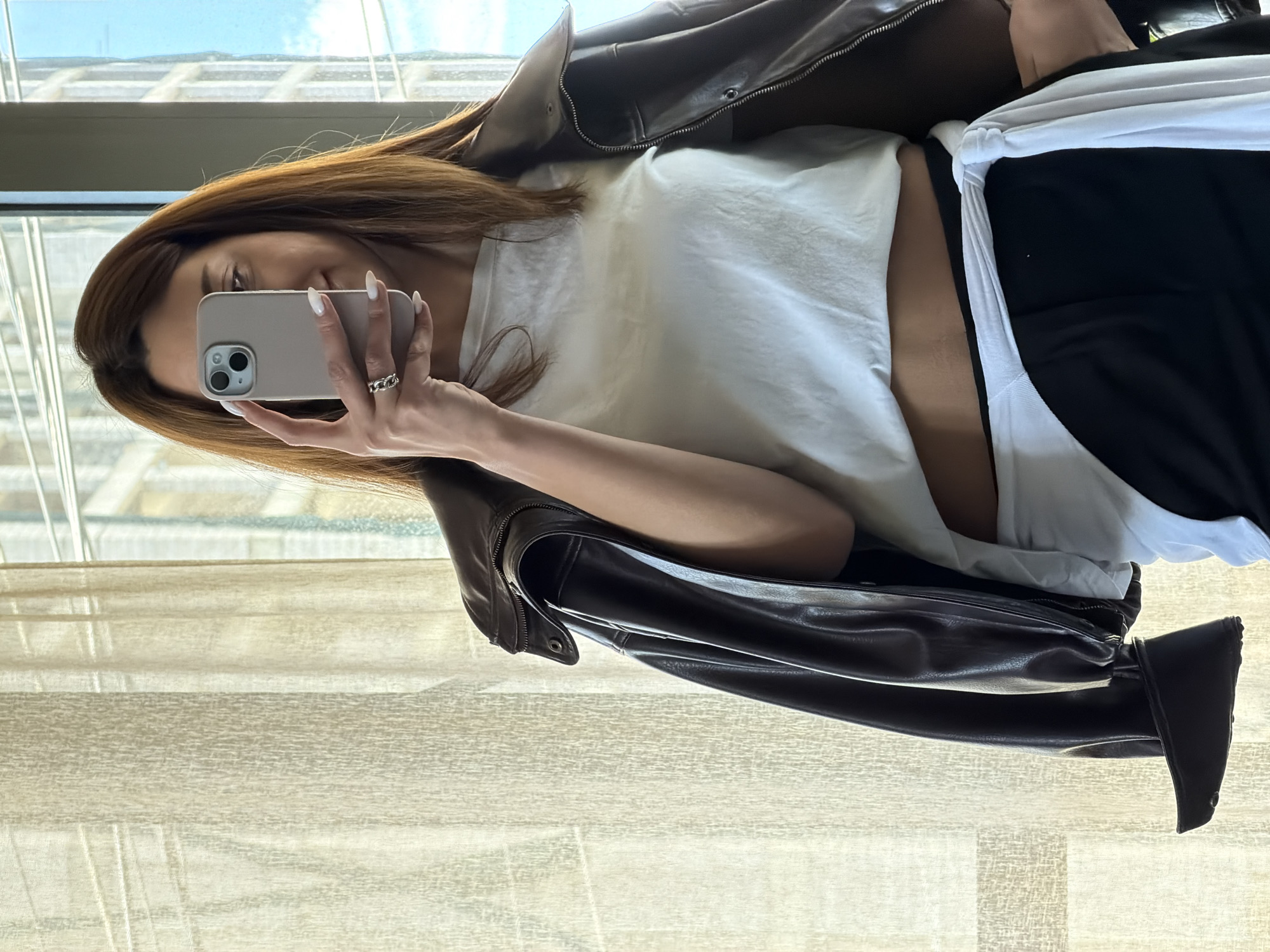The image captures a woman taking a mirror selfie indoors, likely in Yerevan, Armenia. She is seen from the chest up, with her face partially obscured by the phone she is holding. She has long, straight, light brown hair.
She is wearing a dark brown leather jacket over a white, seemingly cropped t-shirt, which reveals a small portion of her midriff. A white garment, possibly another t-shirt or part of the one she is wearing, is tied around her waist over dark, possibly black, bottoms. Her left hand, adorned with a ring featuring interconnected links on her ring finger, holds a light gray or beige-cased iPhone with visible dual cameras. Her nails are long and manicured with a light, possibly white, polish.
The background behind her reveals a large window looking out onto a bright, clear sky and distant buildings, suggesting a city skyline view from a relatively high floor. The light streaming through the window indicates it is daytime. Below the window, a light-colored surface, possibly a wooden floor or a textured wall, is visible. There is no visible text in the image.