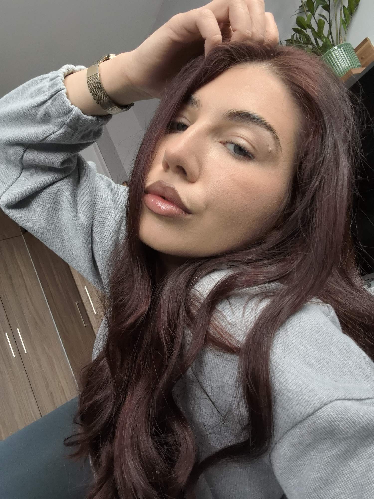 A close-up, slightly high-angle selfie of a fair-skinned woman with long, dark auburn hair. She is wearing a light grey sweatshirt and a gold-toned wristwatch. Her left hand is raised and resting on her forehead. She has full lips and a mole above her left eyebrow. In the background, there is a light brown wooden cabinet and a plant in a green pot on a shelf. There is no information to place the location in Stara Zagora, Bulgaria.