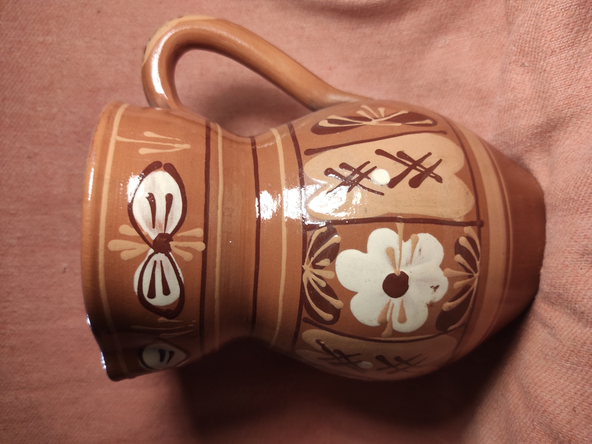 A hand-painted, glazed ceramic pitcher with a handle sits on a textured pink fabric. The pitcher is a warm brown color with lighter tan and dark brown painted floral and bow designs. The pitcher is empty and upright, with its handle curved upwards. The background fabric is draped, creating soft folds. The scene is evenly lit, casting gentle shadows. The location is not discernible from the image.