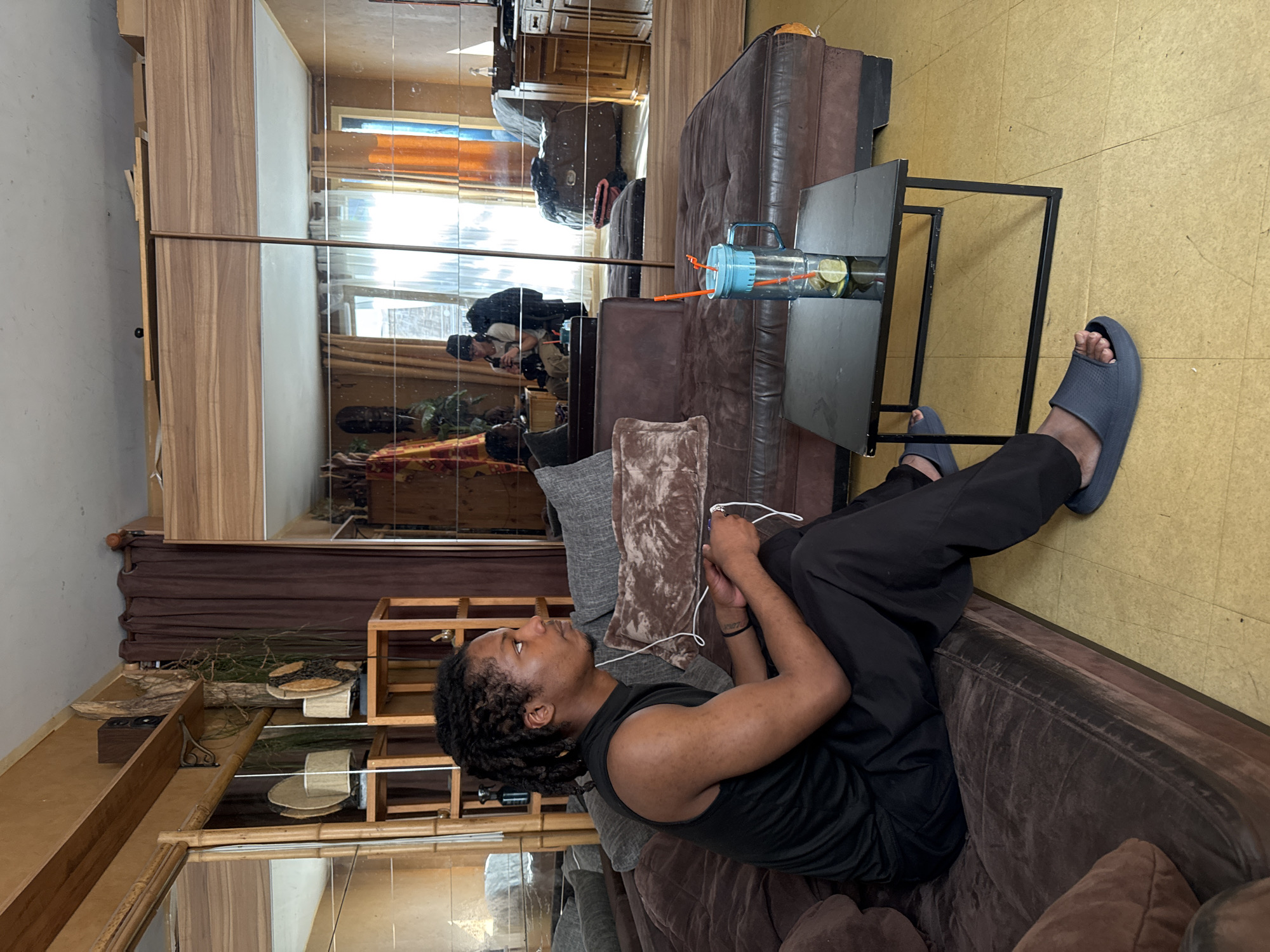The image shows a man relaxing on a brown, suede-like sofa in what appears to be a living room or common area. The man, who has short dreadlocks and is wearing a black sleeveless top, dark pants, and gray slide sandals, is lying on his left side. He holds white wired earbuds, suggesting he is listening to something or using a connected device. His gaze is directed upwards or forward, indicating a relaxed posture.
Next to the sofa, a small black metal side table holds a transparent blue water bottle filled with water and citrus slices (likely lime or lemon), with an orange straw protruding. The floor is a light yellow or beige color, possibly tiled or linoleum.
A significant portion of the room's wall opposite the sofa is covered in mirrors, creating multiple reflections. These reflections show the person taking the photograph, visible with a camera or phone, wearing a dark top and a cap. The mirrors also reflect natural daylight coming through windows, indicating it is daytime. Further reflections reveal parts of a room that might be a kitchen or another area with wooden cabinetry and shelves, as well as decorative items like dried branches and wooden containers. A wooden shelf unit is directly visible near the man's head, adorned with similar natural elements.
The scene conveys a casual and relaxed indoor environment. No discernible text is visible within the image. The given location context is Orsay, France.