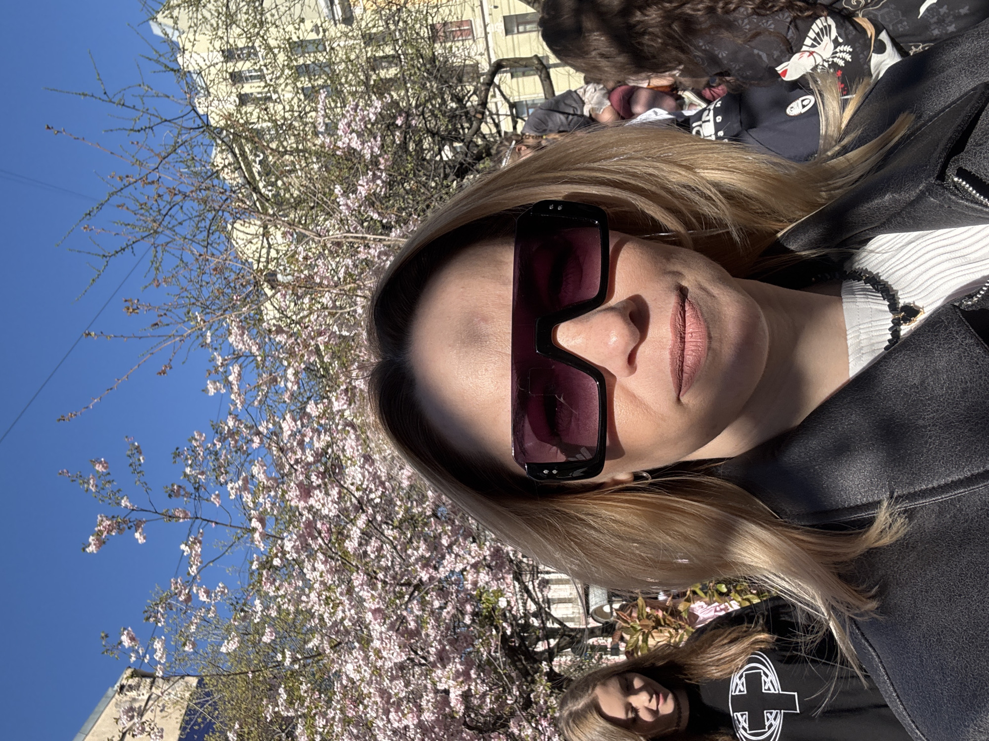 A medium shot captures a woman wearing oversized sunglasses and a black leather jacket. Her blond hair is highlighted by sunlight. Behind her, there is a blooming tree with pink flowers and a building. Another person is partially visible in the bottom right corner of the frame wearing a black t-shirt with a white cross design. The setting is likely Saint Petersburg, Russia, based on architectural style.