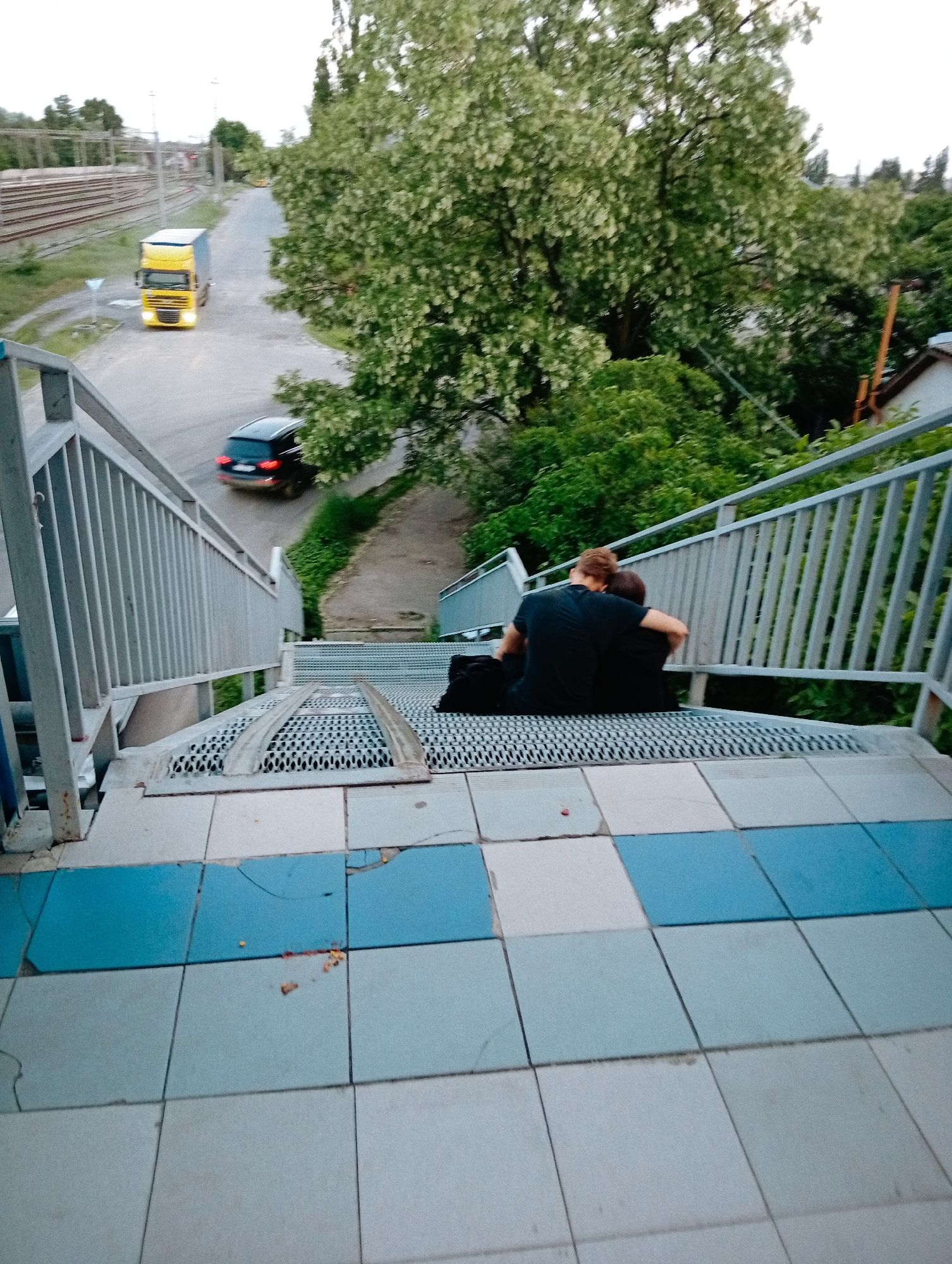 The image shows two people sitting on metal stairs with blue and gray tiled landings. They are embracing, both wearing dark shirts. The stairs appear to be part of an elevated pedestrian walkway. In the background, a road is visible with a yellow truck and a dark-colored car. To the left of the road are train tracks. Lush green trees and other vegetation are present. The photo was taken in Poltava, Ukraine.