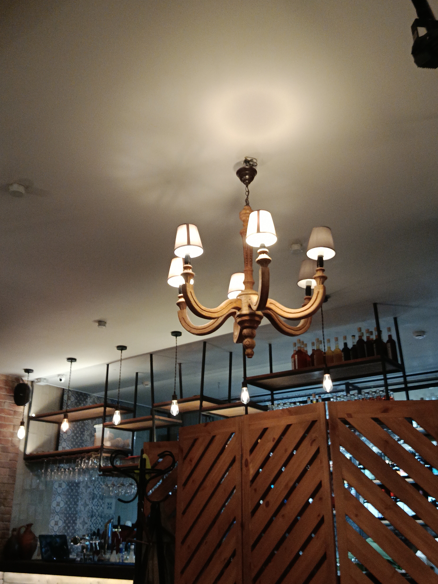 The image shows the interior of a bar or restaurant in Moscow, Russia. A wooden chandelier with multiple lights hangs from the ceiling. The ceiling is light-colored and has multiple light fixtures. In the background, there is a bar area with shelves holding bottles and glasses. A wooden lattice screen partially obscures the view of the bar. The lighting in the scene is warm and inviting.