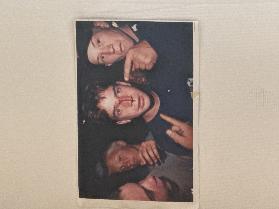 This image is a physical color photograph, adhered to a white, textured surface, likely a wall, with a small piece of light-colored tape visible at the bottom center of the photo. The photograph itself depicts five young males in a dimly lit, interior setting, which is not otherwise identifiable.
In the center, a young male with short, dark, wavy hair and a dark t-shirt is visible. A prominent red mark, consistent with dried blood, forms a cross shape on the bridge of his nose and extends towards his upper lip. His left hand is raised in a gesture with the index and pinky fingers extended. To his immediate right, another young male, wearing a dark cap and a dark shirt with a visible insignia on his right shoulder/chest area, points his right index finger at the central male's nose. His expression is neutral. A third male's face is partially visible on the far left, looking towards the central figure. In the background, to the left of the central figure, a fourth male with light-colored hair holds a clear glass or cup. A fifth, blurry face is faintly discernible in the dark background to the right of the central figure. The specific location within Paris, France, cannot be determined from the visual information present in the image.