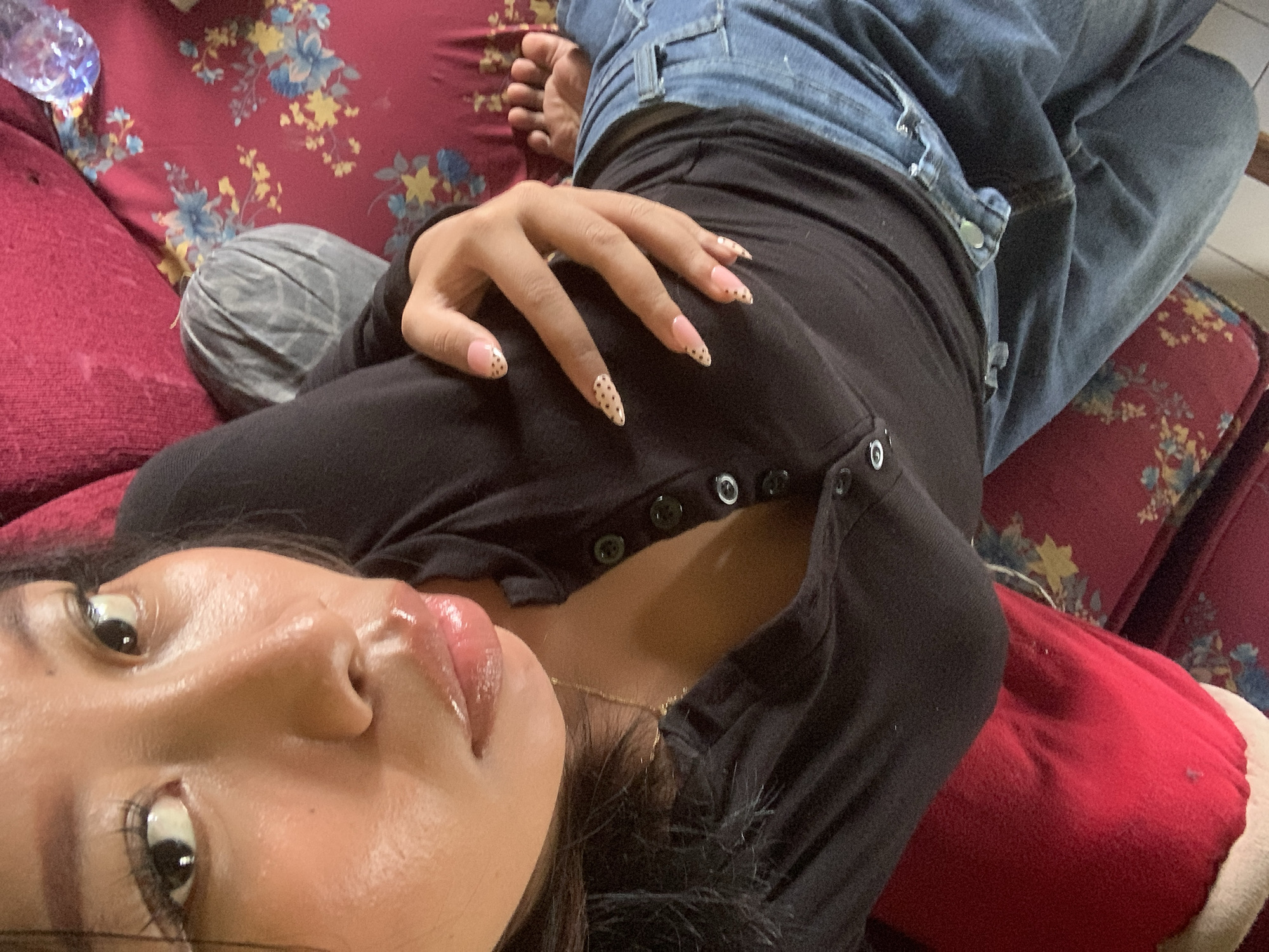 The image shows a close-up, overhead view of a person lying down on a red floral patterned sofa. The person has dark hair and is wearing a black long-sleeved shirt with buttons down the front and blue jeans. Their left hand is resting on their chest, displaying nails painted with a light pink base and small brown dots. The person's face is partially visible, with their eyes looking towards the camera. Their lips are glossy and appear to be slightly parted. To the upper right of the image, a partially visible clear plastic bottle with a blue cap is on the sofa. The overall setting is an indoor environment, consistent with a living room or lounge area. The image does not provide any specific information or visual cues to determine a location within the city of Jajag, Indonesia.