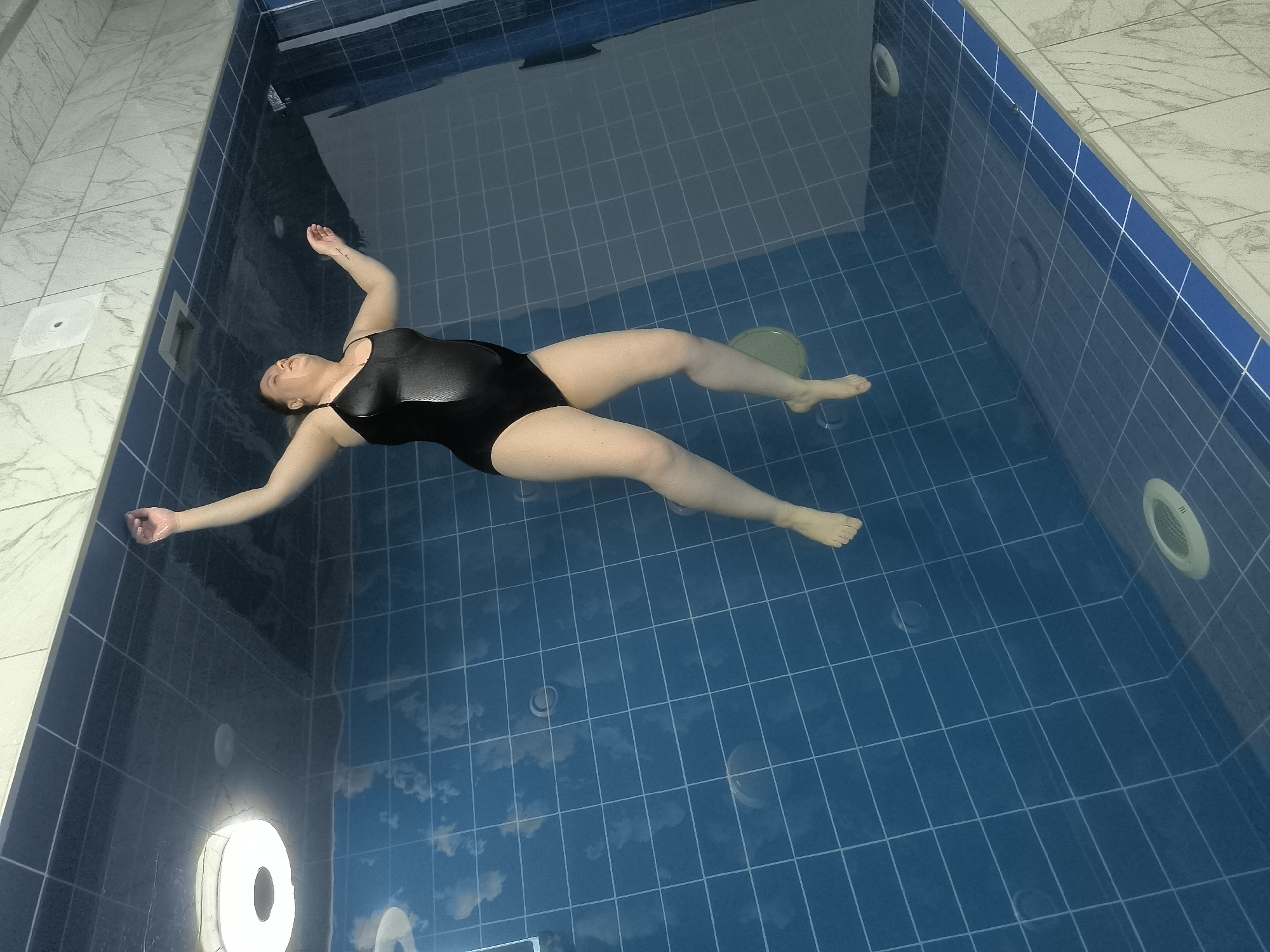 A person is floating on their back in a tiled swimming pool. The person is wearing a black swimsuit and their arms are outstretched to the sides. The pool is filled with blue water, and the bottom of the pool is tiled with a grid pattern. There are lights on the walls of the pool, and a white drain is visible on the left side of the image. The surroundings outside the pool are tiled with white and grey marble-like tiles. The image appears to be taken from above, looking down into the pool. It is impossible to determine the location in the city of Kashino, Russia from the image.