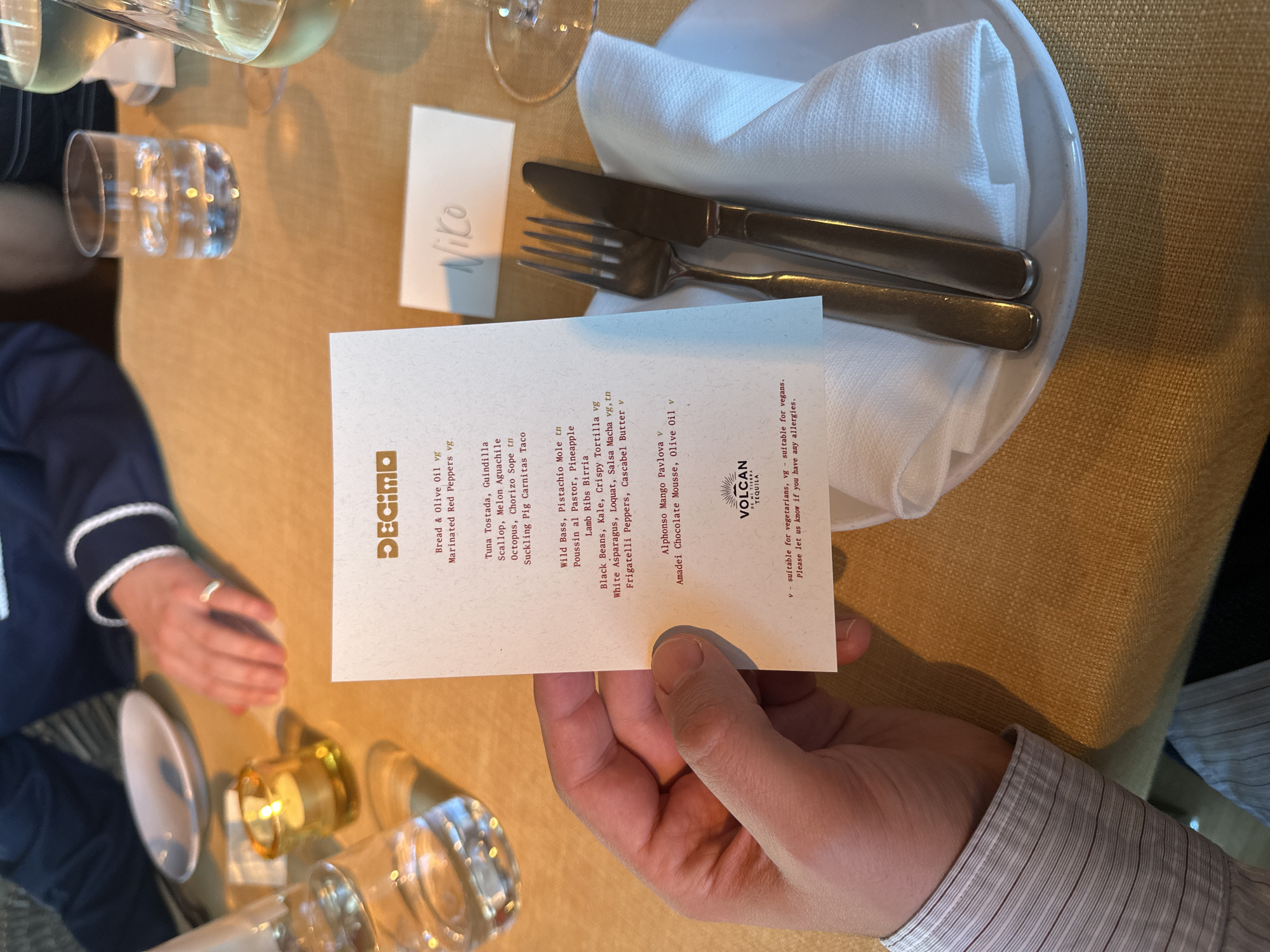 The scene captures a dining experience at the restaurant Decimo in London, United Kingdom. A diner's hand holds the restaurant's menu, showcasing a selection of dishes, including "Bread & Olive Oil," "Tuna Tostada," "Wild Bass," and "Amadei Chocolate Mousse." The menu also indicates vegetarian and vegan options with "v" and "vg" designations. Next to the menu is a place card with the name "Niko" written on it. The table setting includes a white plate with silverware, a water glass, and what appears to be a candle. The table is covered with a beige cloth. In the periphery, parts of two other diners are visible, one in a navy and white top, and another in a striped dress shirt.