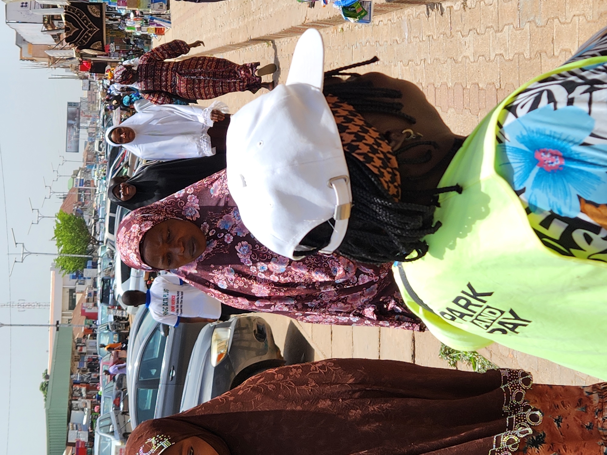 The image depicts a sunny outdoor scene, potentially a marketplace in an urban environment. People wearing traditional Islamic attire are prominently featured; several women wear hijabs in various colors and patterns. There's a person in the foreground wearing a bright yellow vest. A line of parked cars is visible in the background, indicating a busy street. The scene suggests a vibrant and bustling community area.