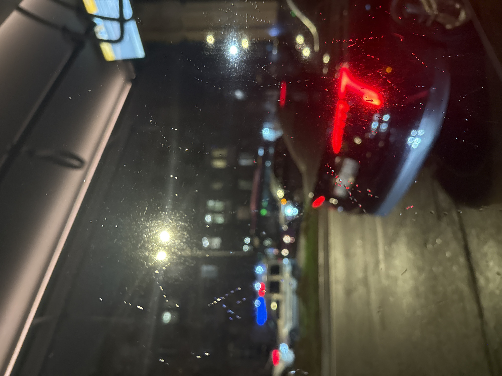 The image depicts a view through a rain-streaked window at night. Outlined through the glass, it seems to be a street scene, probably captured from a vehicle. Outside, a row of streetlights cast a bright glow. The taillights of a dark-colored car can be seen in the close foreground to the right, appearing to be at a standstill. Across the street, several buildings are visible, punctuated by numerous lights, suggesting an urban setting. Based on the urban landscape, it is reasonable to assume this location is in the city of Yerevan, Armenia.