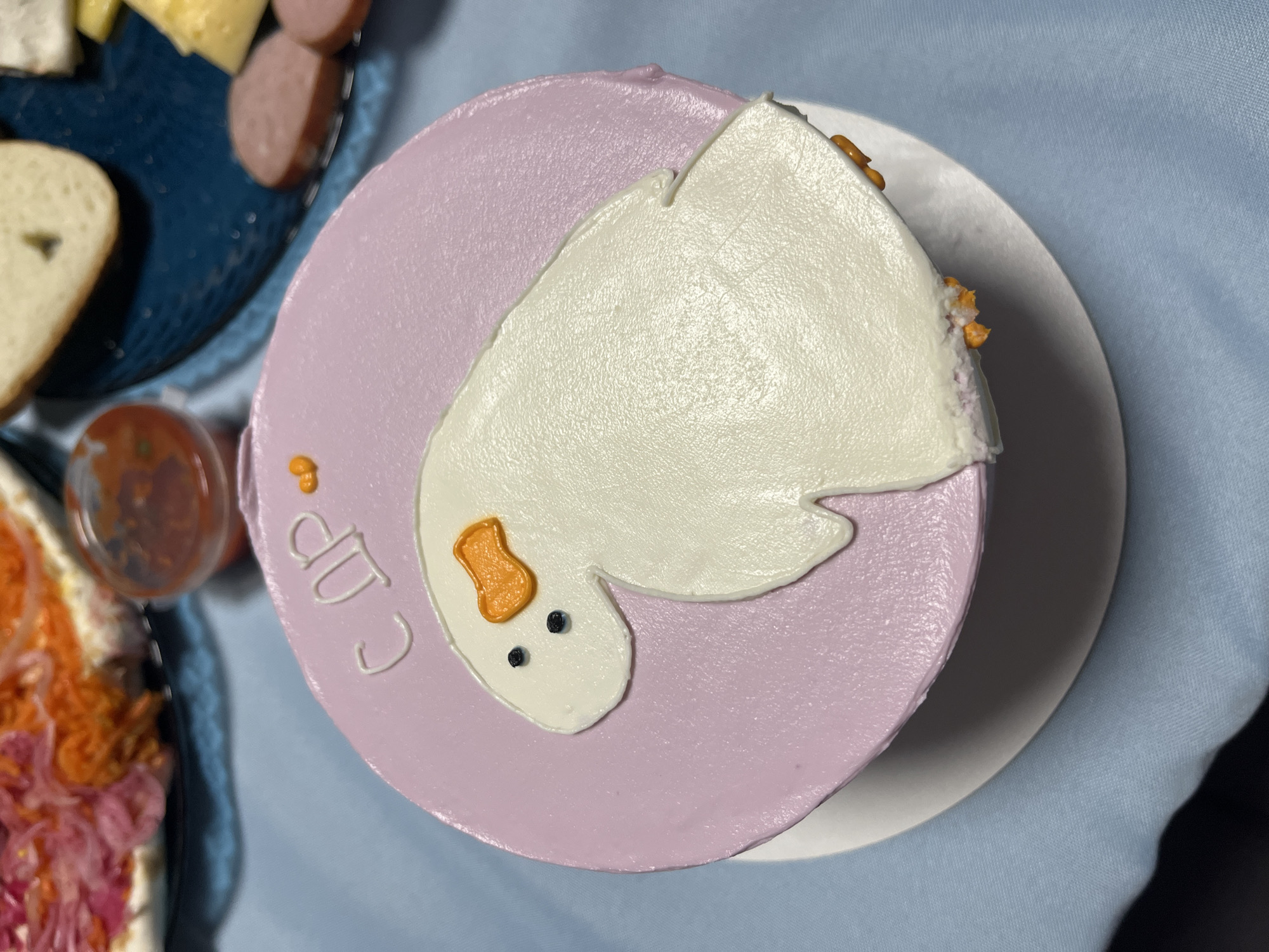 A top-down shot shows a round cake on a white plate, set on a light blue tablecloth. The cake has a light purple frosting base, and a white frosting design resembling a goose with an orange beak and black eyes. The words "Cup" are written in white frosting along the side of the cake.
To the left of the cake, partially visible, are components of a charcuterie board. A dark blue dish holds sliced sausage, cheese, and bread. A small plastic container filled with red sauce is also visible. Additionally, another dish with a salad containing red cabbage is partially in view.
The location cannot be identified from the image.