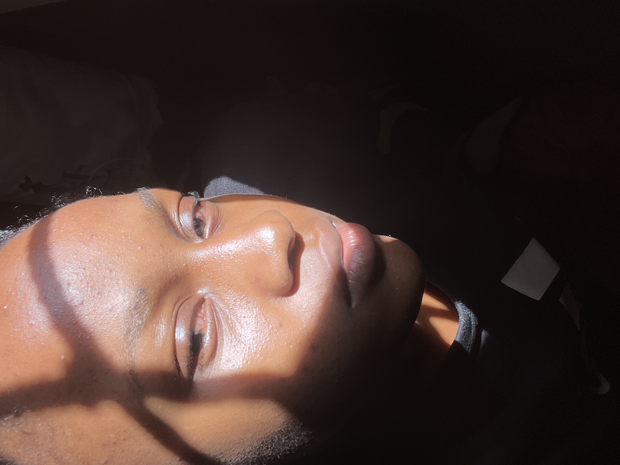 A close-up, low-angle shot captures a person's face illuminated by dramatic sunlight casting a shadow across their forehead and eye. The subject's skin appears to have a dewy sheen. The background is dark, obscuring details of the environment. The image is cropped to focus on the subject's facial features. There is no discernible indication of the location being Rafin Par, Nigeria.