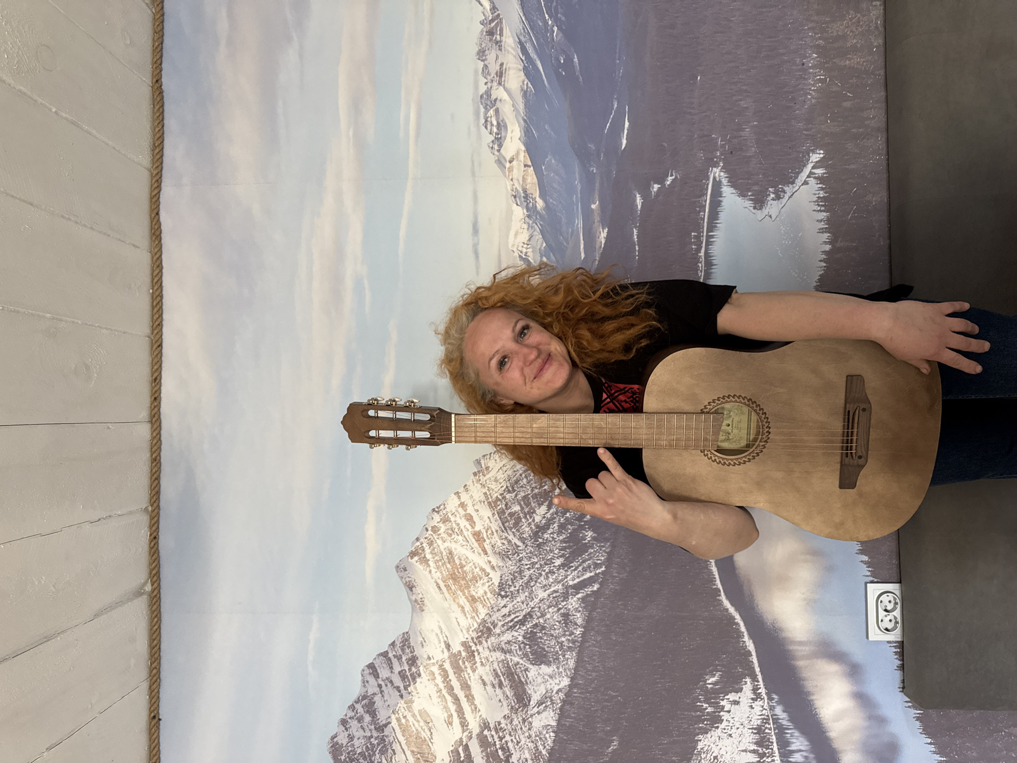 The media file displays an image rotated 90 degrees counter-clockwise, showing a woman with an acoustic guitar against a wall featuring a mountain mural. The woman has long, curly, reddish-blonde hair and is smiling, looking towards the viewer. She is wearing a black t-shirt with a red pattern visible on the shoulder/chest area and dark blue trousers. She is holding a light-colored acoustic guitar; her left hand makes a "rock on" gesture near the guitar's body, while her right arm is draped over the instrument.
The background is dominated by a large wall poster or mural depicting a snowy mountain range under a cloudy sky. The mountains are mostly grey and white, with some lighter patches suggesting sunlight. To the left side of the image, there is a section of a white, horizontally-paneled wooden wall, separated from the mountain mural by a vertical rope. On the lower right, partially visible behind the woman and guitar, is a dark grey surface, and an electrical outlet with two circular sockets is embedded in the mountain mural near the bottom edge.
The guitar features a dark brown fretboard and headstock with metal tuning pegs. A decorative rosette around the soundhole has faint, unreadable text, possibly a brand name like "ANTONIO." The woman's expression is joyful and playful. The setting appears to be an indoor space, possibly themed, located in Chaikovsky, Russia.