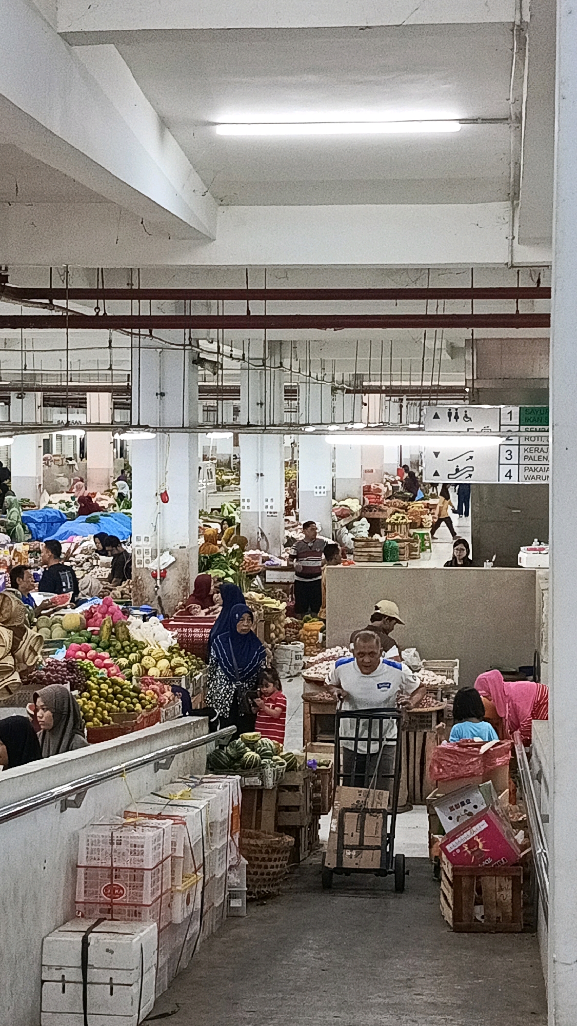 The image depicts an indoor market scene in Nologaten, Indonesia. The market appears to be bustling with activity, featuring stalls overflowing with a variety of fresh produce, including fruits and vegetables. Several individuals are visible, some attending to the stalls and others browsing or moving through the market.
In the foreground, a man is pushing a trolley laden with boxes, heading towards the right side of the frame. To his left, a young girl in a striped red shirt is looking down at a mobile device, while an adult woman in a blue hijab stands nearby. Behind them, there are various crates and baskets containing produce.
Further into the market, multiple vendors and shoppers are present. One stall prominently displays a colorful assortment of fruits such as oranges and what appear to be mangoes or similar large fruits. Another stall has a woman in a pink headscarf and an adjacent stall with a person wearing a blue shirt. The market has a concrete floor and is illuminated by fluorescent lighting fixtures mounted on the ceiling. Structural elements like beams and columns are visible, indicating an indoor, possibly covered, market setting. Signs are visible in the background, one of which appears to be a directional or informational sign with numbers and text in a foreign language. The overall impression is of a vibrant and active marketplace.