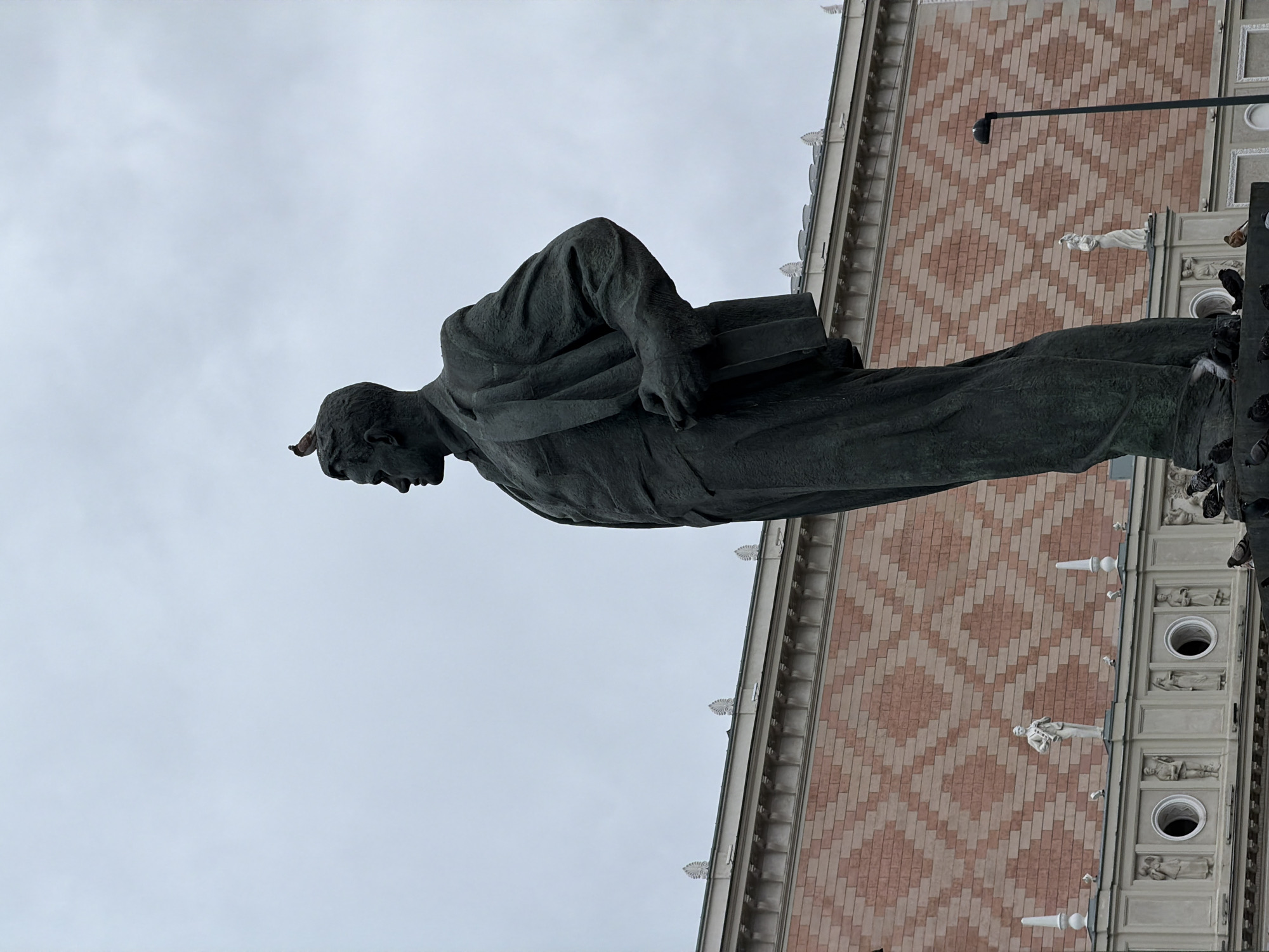 A low-angle shot of a statue of a man in front of a building in Moscow, Russia. The man is standing and has his head turned downward. He is wearing a suit and carrying a book in his left hand. The statue is dark grey. The building behind him has a brick patterned facade with a white base and several small statues along the top. The sky is light grey. There are birds on the statue's base.