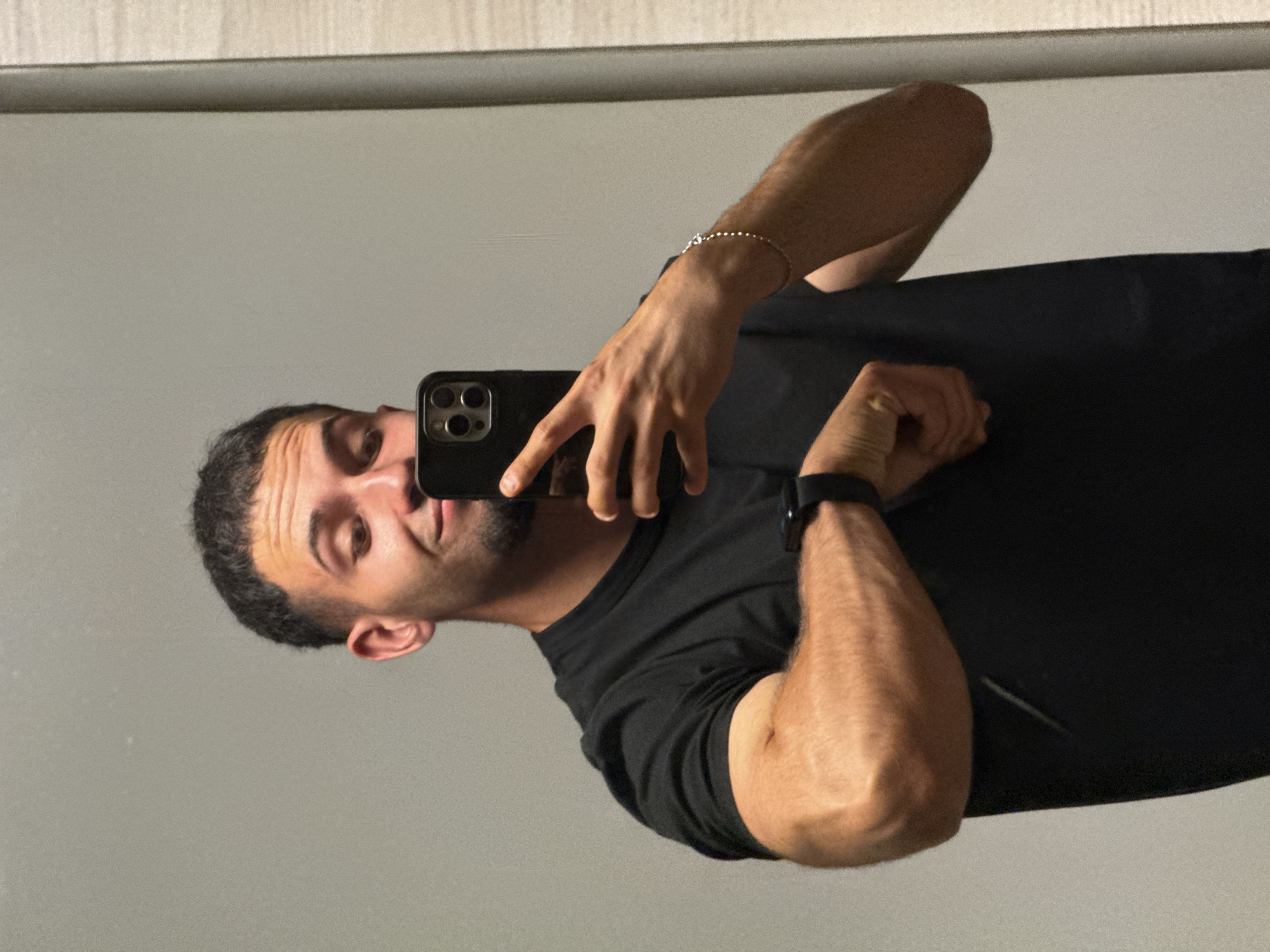 A man is taking a mirror selfie in a room. He is wearing a black t-shirt and a smartwatch. He is holding a black smartphone with a black case in his right hand, and his left hand is resting on his arm, showing off his muscles. He has short, dark hair and a beard. The room has light gray walls and a vertical beige element on the right side, which could be a door or a wall panel. The lighting is neutral, and there are no other people or objects visible in the frame. The image does not contain any specific details that would allow for its localization in Shumen, Bulgaria.