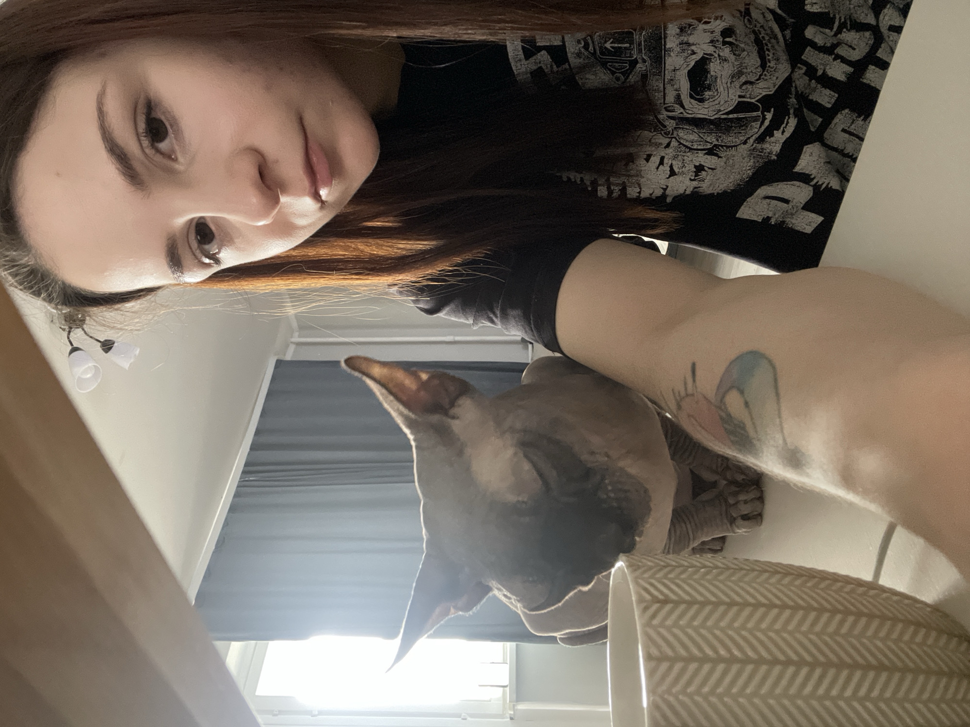 An overhead shot captures a young adult woman and a Sphynx cat indoors. The woman, with long dark brown hair, is positioned in the upper left, looking directly towards the camera. She has a neutral expression and is wearing a black t-shirt featuring a partially visible white graphic print, which appears to depict a skull or monstrous face and some stylized text, possibly "DUTCH". A colorful tattoo, featuring blue, pink, and orange tones in an abstract or bird-like design, is visible on her right upper arm.
To the right and slightly below the woman, a greyish-brown Sphynx cat is seen with its large ears prominent, looking downwards or away from the camera. The cat's wrinkled skin is visible. In the foreground on the right, a beige ceramic cup or planter with a white chevron or herringbone pattern is partially visible on a white table or surface.
The background shows a window with light-colored blinds or curtains, allowing natural daylight to illuminate the scene, suggesting it is daytime. A small light fixture with two white, bell-shaped shades hangs subtly in the upper left background. Another flat, brown surface, possibly a box or bag, is on the far left. The overall setting appears to be a casual indoor space.