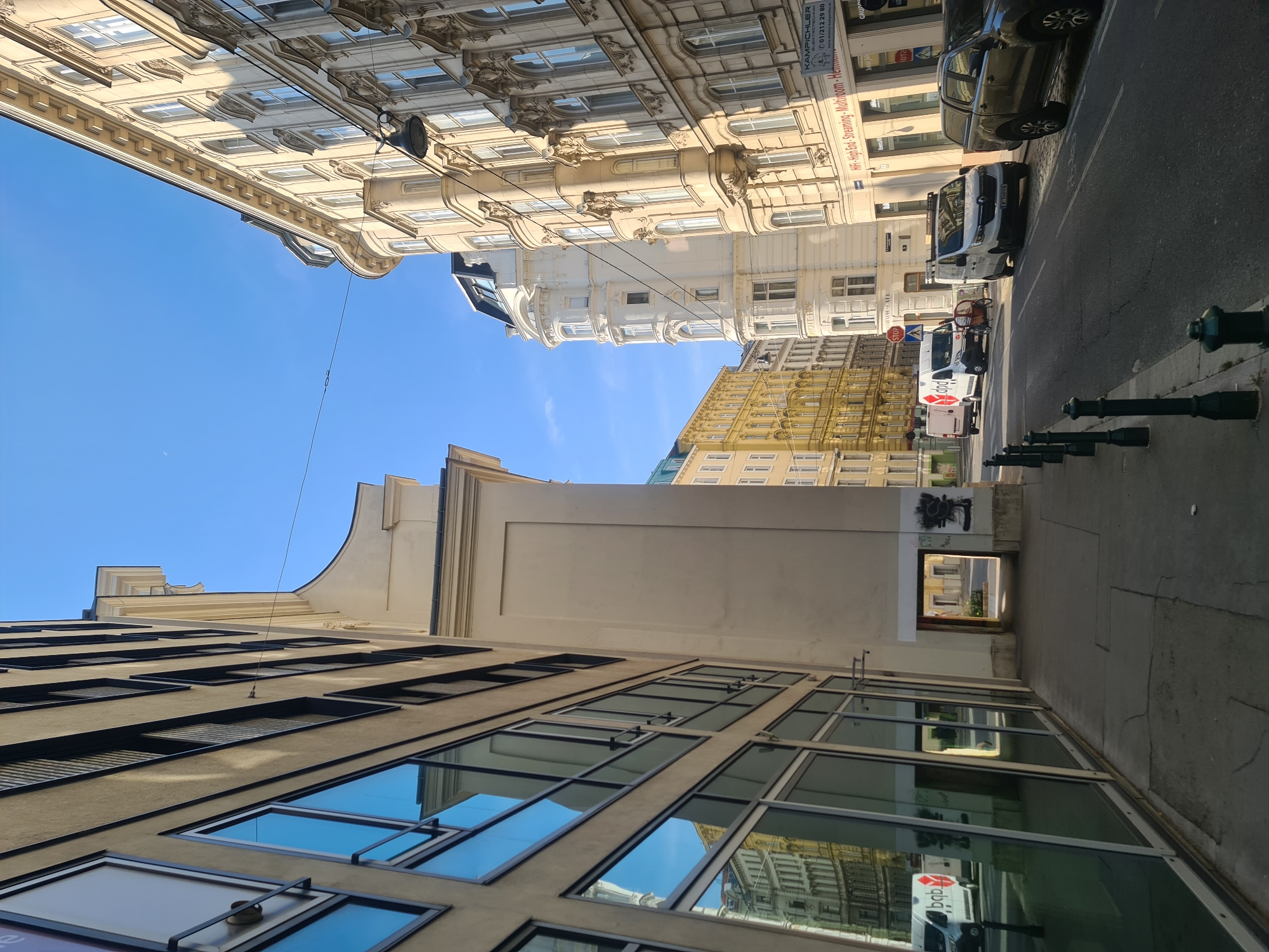 This is a daytime, eye-level, medium shot of a street in Vienna, Austria. The street is lined with buildings of varying architectural styles and heights. On the left, a modern building with large, reflective windows dominates the foreground. The windows reflect other buildings across the street, including those with intricate classical detailing.
The street itself is paved with asphalt and marked with white lines. A sidewalk runs along the left side of the street, made of concrete slabs. Several black bollards are spaced along the edge of the sidewalk.
Several vehicles are parked or driving on the street. These include a white DPD delivery van, a white Renault van with a bicycle rack on the back, another white van, and a dark-colored pickup truck.
On the right side of the street, older buildings with ornate facades, arched windows, and decorative elements are visible. One building has a sign that reads "KAMPICHLER" and advertises "Hi-Fi - High End - Streaming". Another building in the background is painted a pale yellow. Further down the street, a white, multi-story building with rounded architectural features stands out.
The sky is clear and blue. The overall impression is of a bustling, urban environment with a mix of historical and modern architecture.