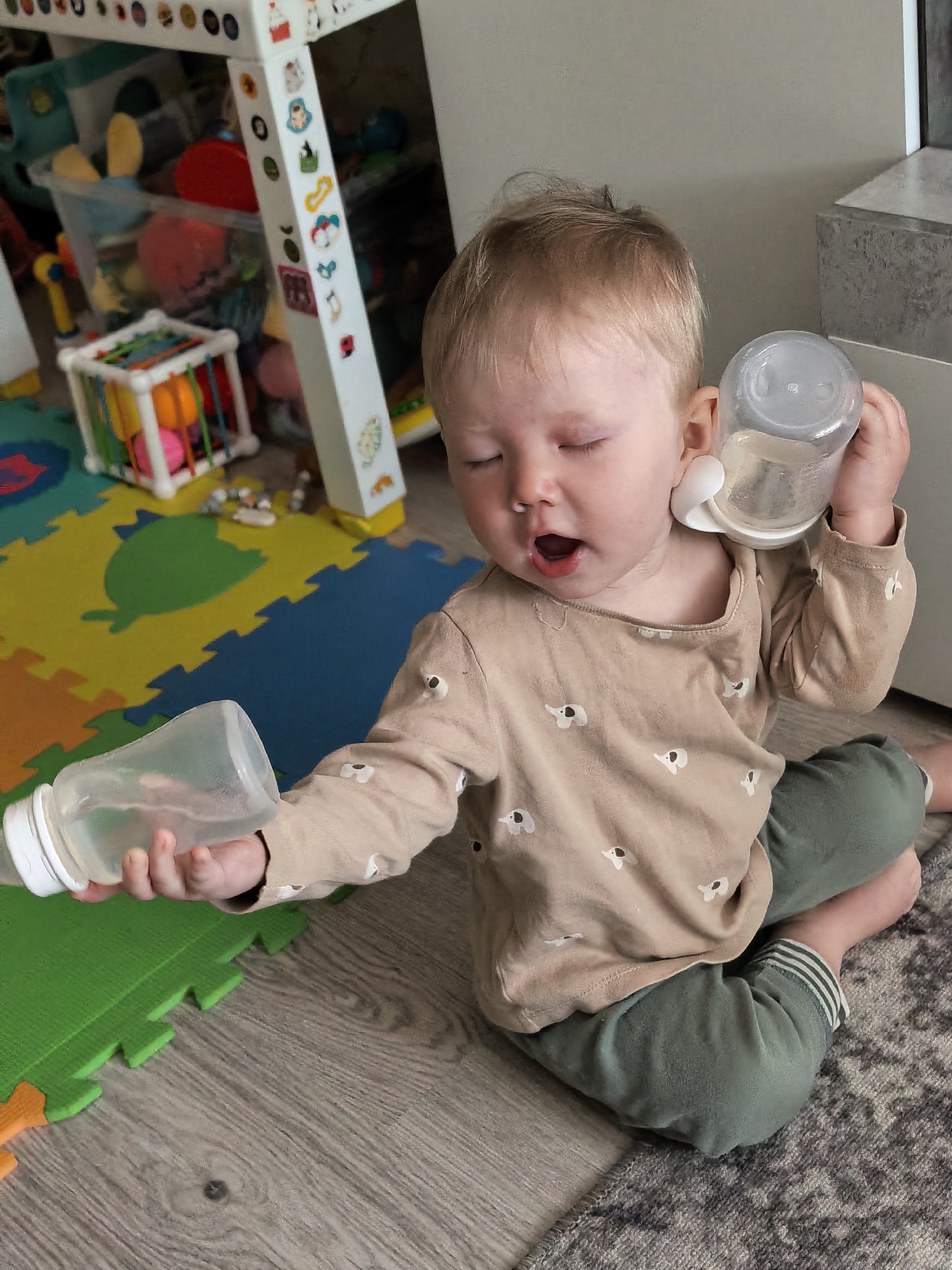 A young child with blond hair is sitting on the floor. They are wearing a light brown long-sleeved shirt with small white elephant patterns and green pants. The child is holding two baby bottles, one to their ear like a phone and the other in their hand. The child's mouth is open and their eyes are closed. A colorful playmat is visible on the floor near the child, as well as toys and furniture in the background. There is no information available to confirm the location as Sterlitamak, Russia.
