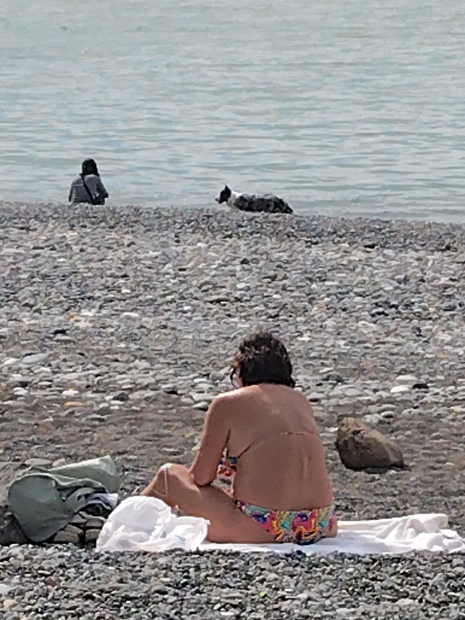 The image depicts a beach scene, likely in Batumi, Georgia, given the presence of a pebble beach along the Black Sea. In the foreground, a woman is seated on a white towel placed on the pebble beach. She is wearing a colorful bikini and has her back to the camera. Her belongings, including a green garment and a white hat, are piled next to her. In the midground, two other people are visible in the water. One appears to be seated or wading in the water, and the other is accompanied by a dog. The background consists of the Black Sea and the sky, which appears overcast. The scene is tranquil, with people enjoying the beach despite the lack of clear skies. The environment appears rugged due to the pebble beach.