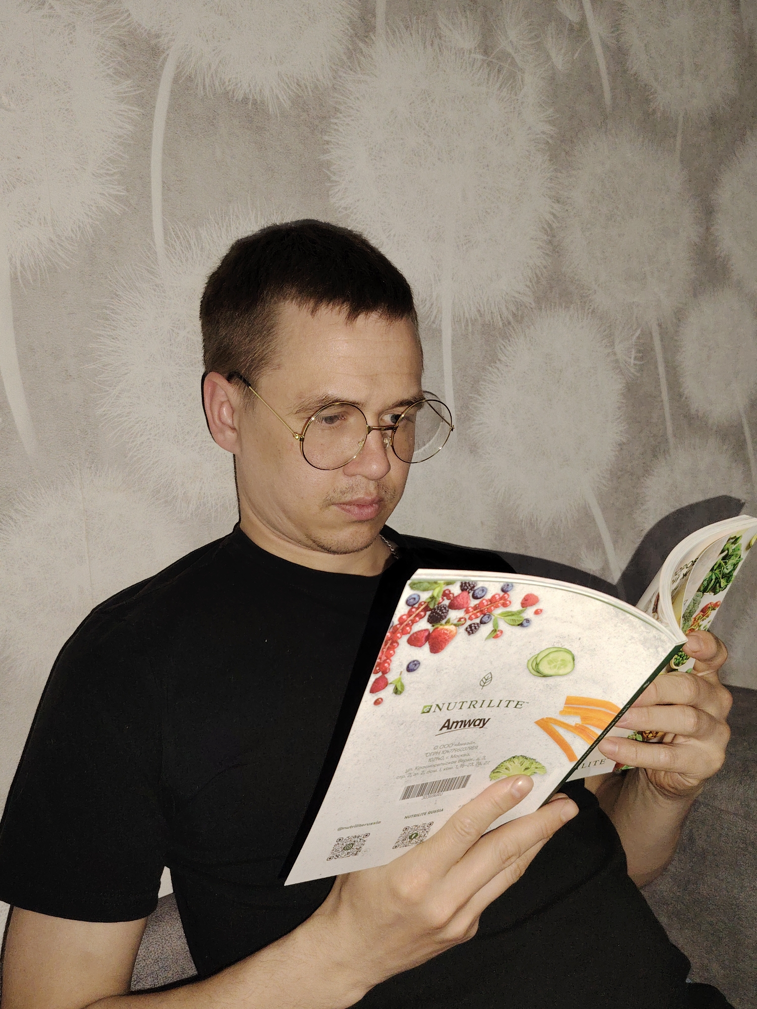 A man with short dark hair and round gold-rimmed glasses sits indoors, holding open a brochure titled "NUTRILITE Amway." The brochure features images of fruits and vegetables. The man wears a black t-shirt. The background is a grey wall with a patterned design. According to the OCR text within the image, the Amway office is located at Ul. Krasnosel'skaya Verki, 3, Moscow, Russia. Chaikovsky, Russia, is not mentioned.