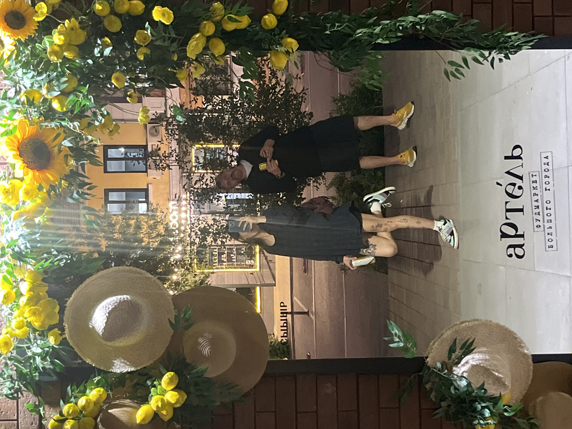 The image captures a reflection in a storefront window at the "Artel" food market, located on Mira Avenue in Krasnoyarsk, Russia. The storefront is adorned with a festive floral arch of sunflowers, lemons, and green foliage.
In the reflection, a woman is seen holding a smartphone, taking a selfie. She is wearing a dark, loose-fitting dress and dark shorts, paired with black and white sneakers. Tattoos are visible on her legs. She is holding a beverage can.
To her right, a man is also visible in the reflection. He has a mustache and is wearing a dark sweater or long-sleeved shirt over a lighter undershirt, and a dark skirt or kilt-like garment. He is holding a beverage can. He is wearing yellow sneakers.
The foreground of the image, outside the reflection, features part of a straw hat decorated with greenery and lemons, positioned on the left side. The floor appears to be paved with light-colored tiles or bricks. The background shows a street scene with buildings and parked vehicles. A sign with Cyrillic text, "А РТЕЛЬ" (Artel) and "ФУДМАРКЕТ БОЛЬШОГО ГОРОДА" (Food Market of the Big City), is prominently displayed at the bottom of the storefront window.