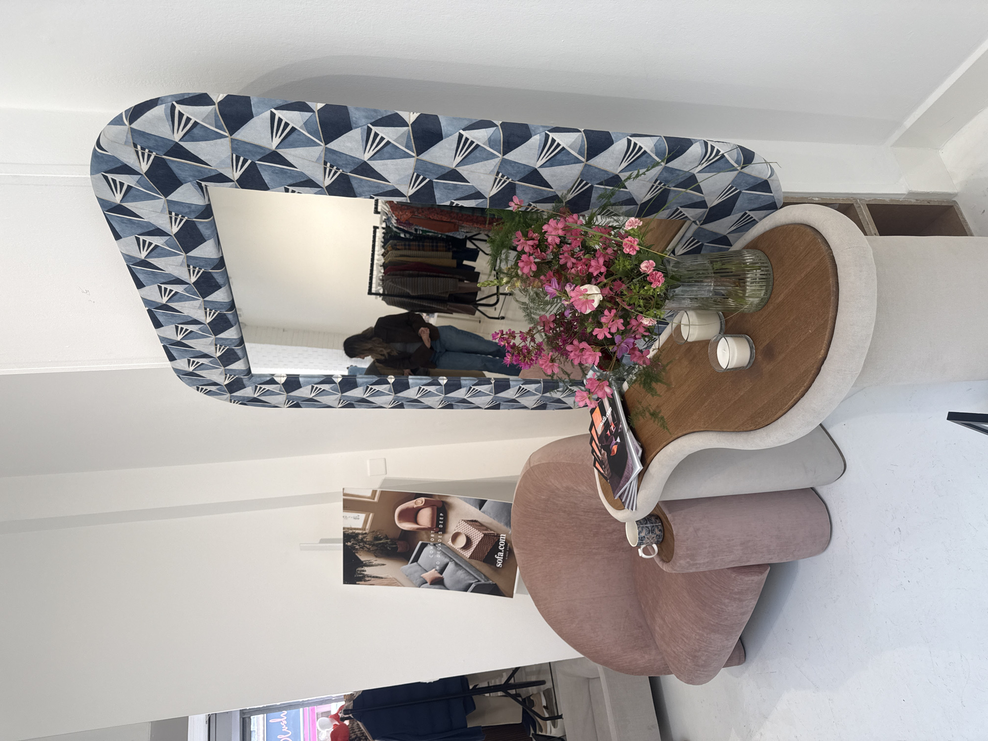 The image captures an interior scene, possibly a store or showroom, in London, United Kingdom. A patterned mirror hangs on a white wall, reflecting a person and a clothing rack. A small table with a wooden top displays flowers in a vase and candles. Next to it, a pink chair is situated. A poster featuring furniture is affixed to the wall. Other clothing can be seen in the bottom part of the image.