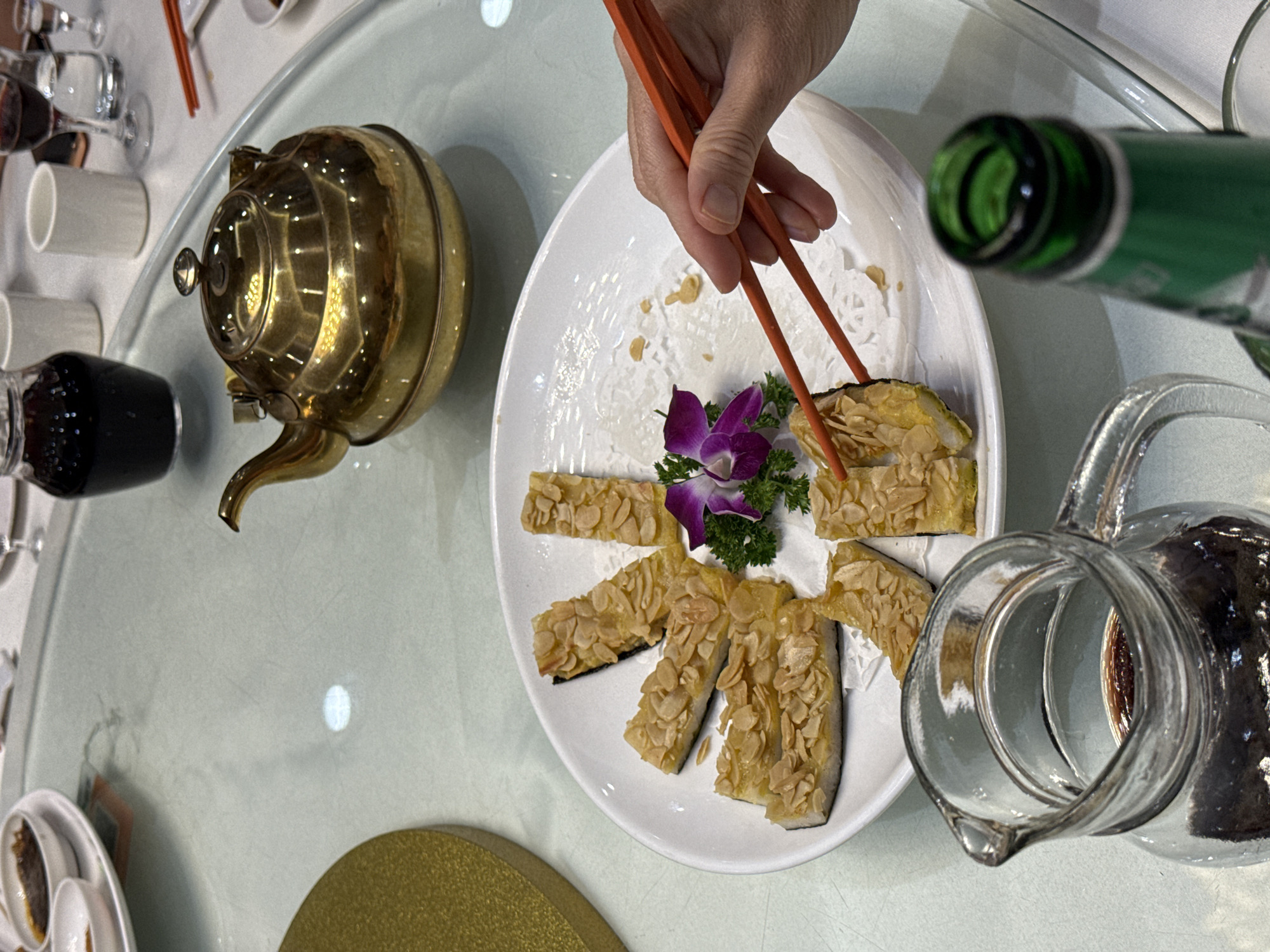 A high-angle shot depicts a dining table scene, likely within a restaurant in the Liwan District, China. Dominating the scene is a white plate, arranged with rectangular slices of food coated with slivered almonds. A small bouquet of purple orchids with green foliage sits in the center of the plate. A hand holds orange chopsticks, about to pick up a slice of food. To the left of the plate, a brass teapot adds a touch of traditional Chinese ambiance. Further to the left are a series of small dishes and glasses. To the right of the plate, there is a glass container with a liquid, along with the green neck of a bottle.