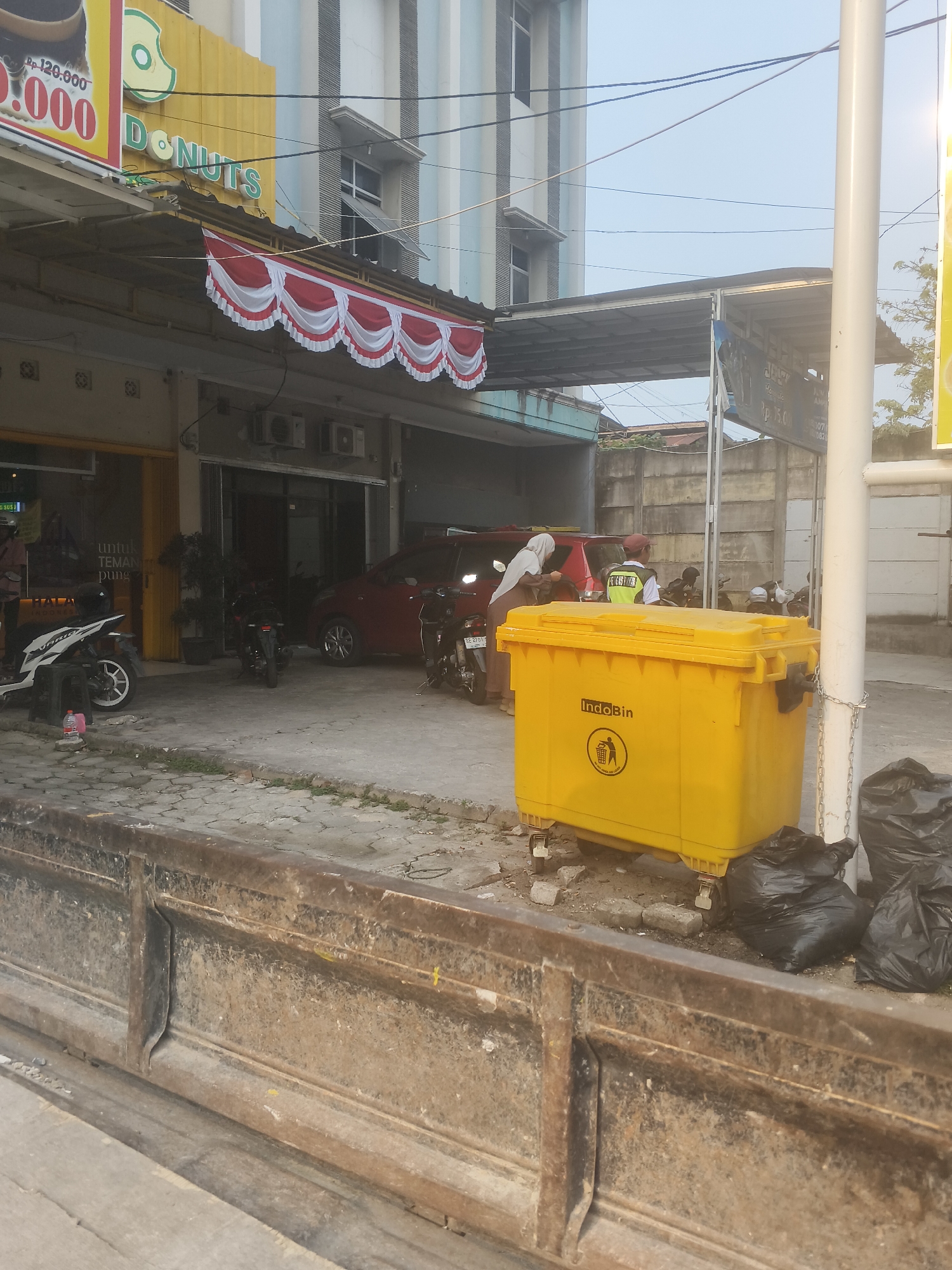 The image shows a street scene in Bandar Lampung, Indonesia. In the foreground, there is a rusty metal barrier. Behind the barrier, a large yellow dumpster labeled "IndoBin" is visible, with black trash bags next to it. A red car and several motorcycles are parked on the street. A building with a yellow facade and "DONUTS" written on it is on the left. A white pole with chains attached to it stands to the right of the dumpster. The sky is clear and bright.