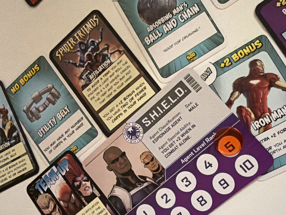 This image presents a close-up, oblique view of multiple overlapping cards from a tabletop game, laid out on a flat, light-colored surface. The game appears to be Marvel Comics-themed.
Prominently displayed is a "S.H.I.E.L.D." character card in the foreground, featuring two male figures: a dark-skinned individual with sunglasses in a blue and white uniform at the top, and a lighter-skinned, blond individual also with sunglasses in a blue top below. The card details "Agent Classification: ESPIONAGE AGENT," "Agent Special Ability: YOU GET +2 WHEN IN COMBAT ALONE," and "Sex: MALE." A vertical "Agent Level Rank" track, numbered 1 through 10, is visible on the left side of this card, with a circular orange marker indicating "5." The word "ALLY" is partially visible to the right.
Above and partially obscured by the "S.H.I.E.L.D." card is a "Utility Belt" item card, depicted with a brown utility belt and the text "YOU MAY HAVE ANY NUMBER OF CARDS IN YOUR HAND." It also shows "NO BONUS" at the top. To its right, the "Spider-Friends" affiliation card features multiple spider-themed characters in action poses, with text including "TO AVOID A TRAP YOU MAY DISCARD A CARD AND ROLL THE DIE. ON A 4 OR HIGHER, CANCEL THE TRAP AND YOU MAY PUT THAT TRAP IN YOUR HAND." and "YOU GET A +2 BONUS WHEN YOU HAVE TWO OR FEWER CARDS IN YOUR HAND."
Further right, partially visible, is the "Absorbing Man's Ball and Chain" card, illustrating a metallic ball and chain, with the descriptive text "GOOD FOR CRUSHING." A blue circle with "500" is visible on this card. Below this, the "Iron Man" character card is partially visible, showing Iron Man in his red and gold suit in a dynamic pose, with a "+2 BONUS" indicator.
To the far left, a "TEAM-UP" card is partially visible, displaying two female characters, one with red hair and another masked. Other card edges and fragments of text are visible in the background, indicating additional game elements. The overall impression is of a game in progress or set up for play.
No visual information within the image indicates a specific geographic location, including whether it is in Tel-Aviv, Israel.