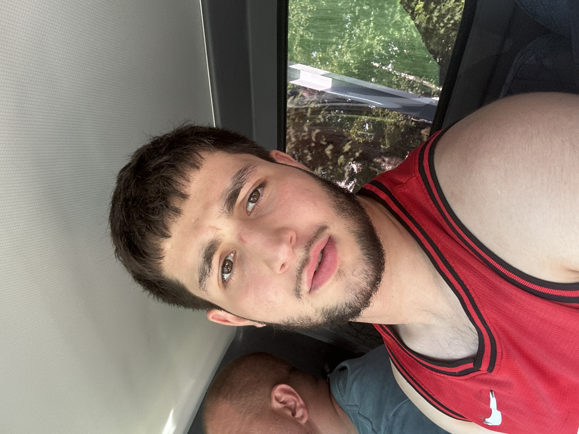 A close-up, selfie-style shot taken inside a vehicle shows the upper body of a young man with short, dark, choppy hair and a light beard. He is wearing a red basketball jersey with black trim and a white Nike swoosh logo. His expression is neutral, with his mouth slightly open. To his left, partially visible, is the bald head and shoulder of another person wearing a teal shirt. The background shows a car interior with a textured ceiling and a window reflecting green trees and foliage, suggesting an outdoor setting. The lighting appears natural, likely from daylight. The image does not contain any identifiable landmarks or elements that would indicate its location within Birobidzhan, Russia.
