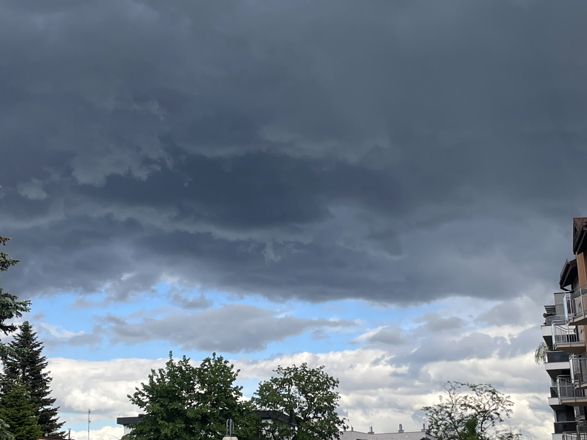 The image shows a sky dominated by a dark, ominous layer of storm clouds. Below this layer, a band of lighter clouds and patches of blue sky can be seen. The lower portion of the image features treetops and the tops of buildings. The buildings appear to be residential, with balconies visible.