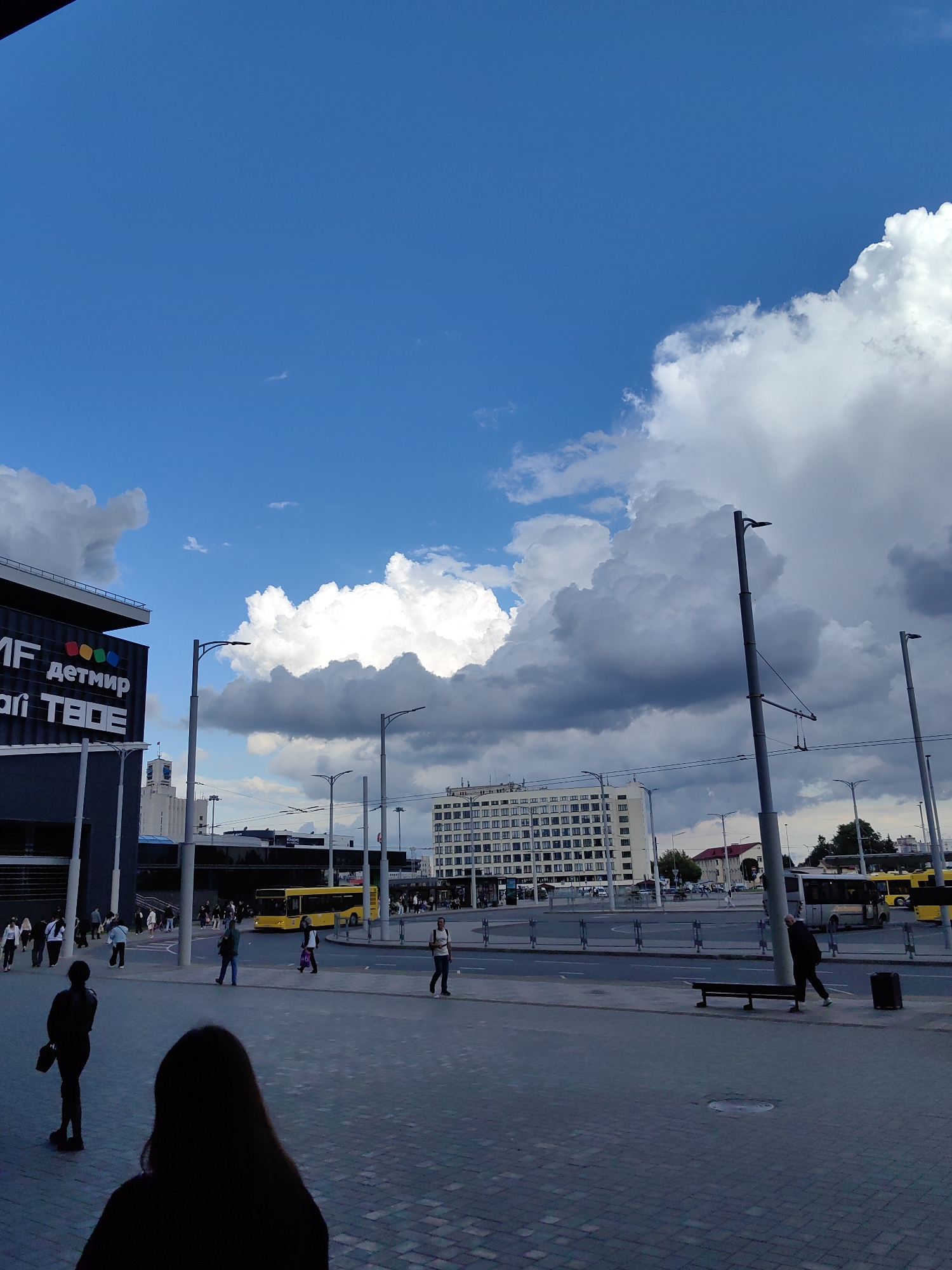 The image depicts an outdoor scene in Minsk, Belarus, likely near the National Library of Belarus, given the architecture and general urban setting. The sky is predominantly blue with large, fluffy white and grey clouds, suggesting variable weather. In the foreground, a paved pedestrian area with a distinct pattern of grey rectangular bricks is visible. Two individuals are in silhouette on the left side of the frame; one facing away from the camera with long hair and the other with their back to the camera, holding a bag. Further back, several people are walking on the pedestrian area and crossing a road.
To the left, a modern building with a dark facade features prominent white lettering that appears to read "MF детмир" and "ari ТВОЕ". This building has a stepped upper section and a railing. Several streetlights on tall poles are strategically placed throughout the scene. Across the road, a large, white, multi-story building with a grid-like pattern of windows dominates the background. Yellow buses are visible on the road in front of this building, indicating a public transportation hub. A park bench and a trash receptacle are present in the mid-ground on the right. The overall atmosphere is that of a bustling urban environment on a day with dramatic cloud formations.