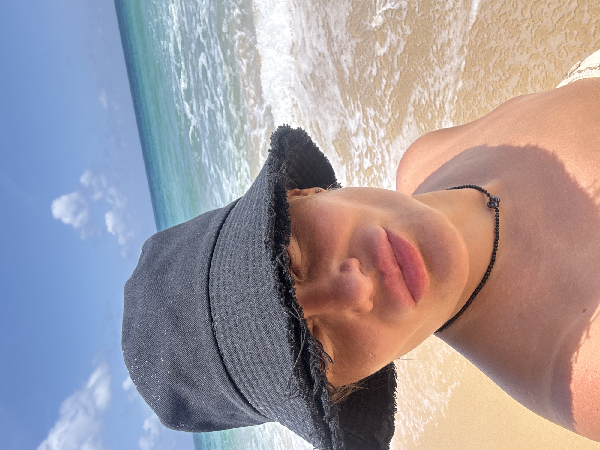 A woman wearing a black bucket hat with frayed edges and a black beaded necklace poses for a selfie on a sandy beach. She is looking upwards, with her eyes mostly closed. The ocean water is a turquoise color with white waves lapping at the shore. The sky is blue with scattered white clouds. The location appears to be a tropical beach, possibly in Choeng Thale, Thailand. The overall lighting suggests it is daytime and sunny.