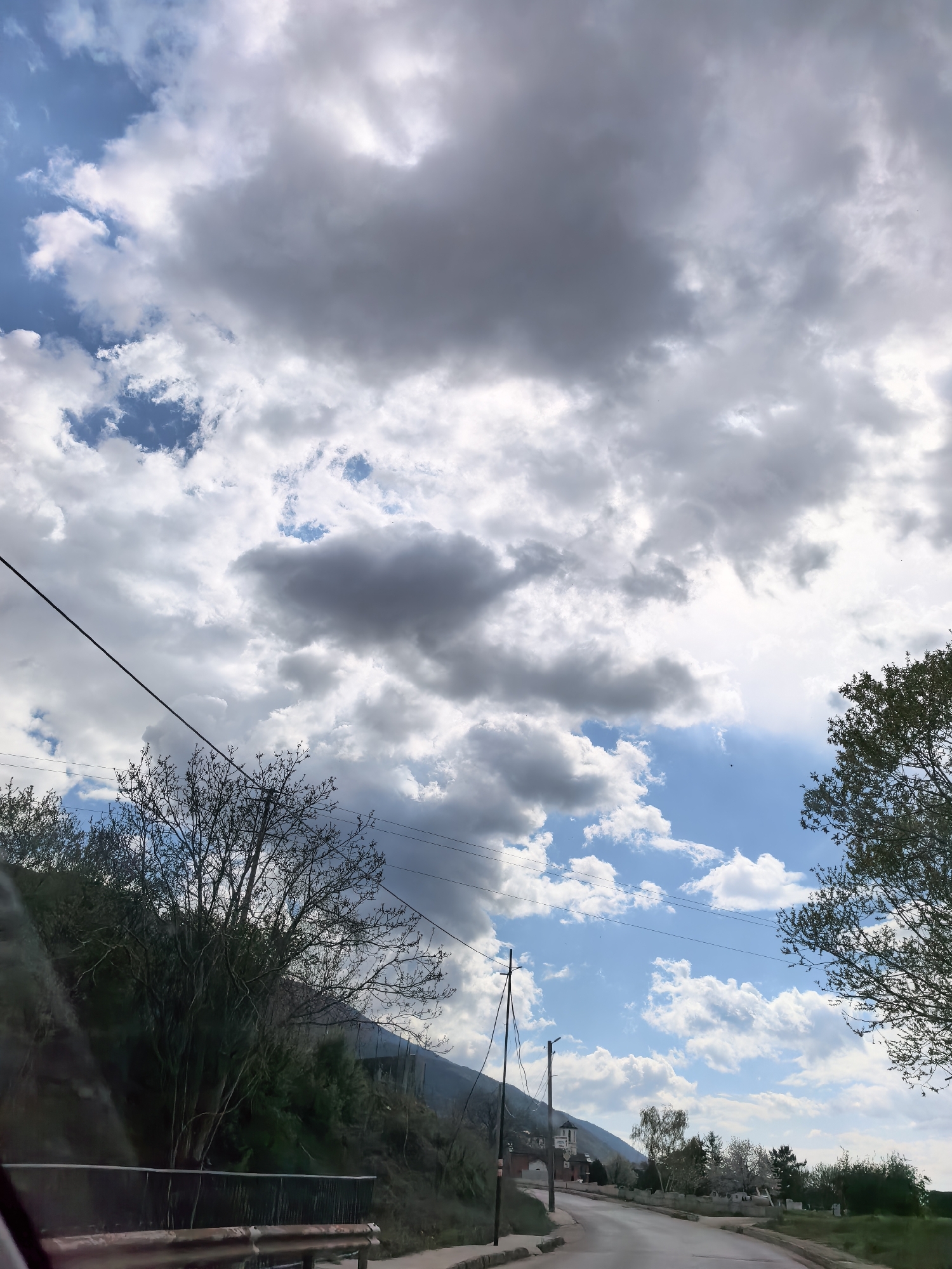 The image shows a street view with a cloudy sky. Power lines stretch across the top portion of the image. Trees are present on either side of the road, with one tree predominantly on the left side and another on the right side. A mountain is seen in the background. The road leads to a village with buildings, including one that looks like a church. A metallic guard rail is visible in the lower left corner, and part of a vehicle dashboard is also visible. The location is Unknown, Unknown.