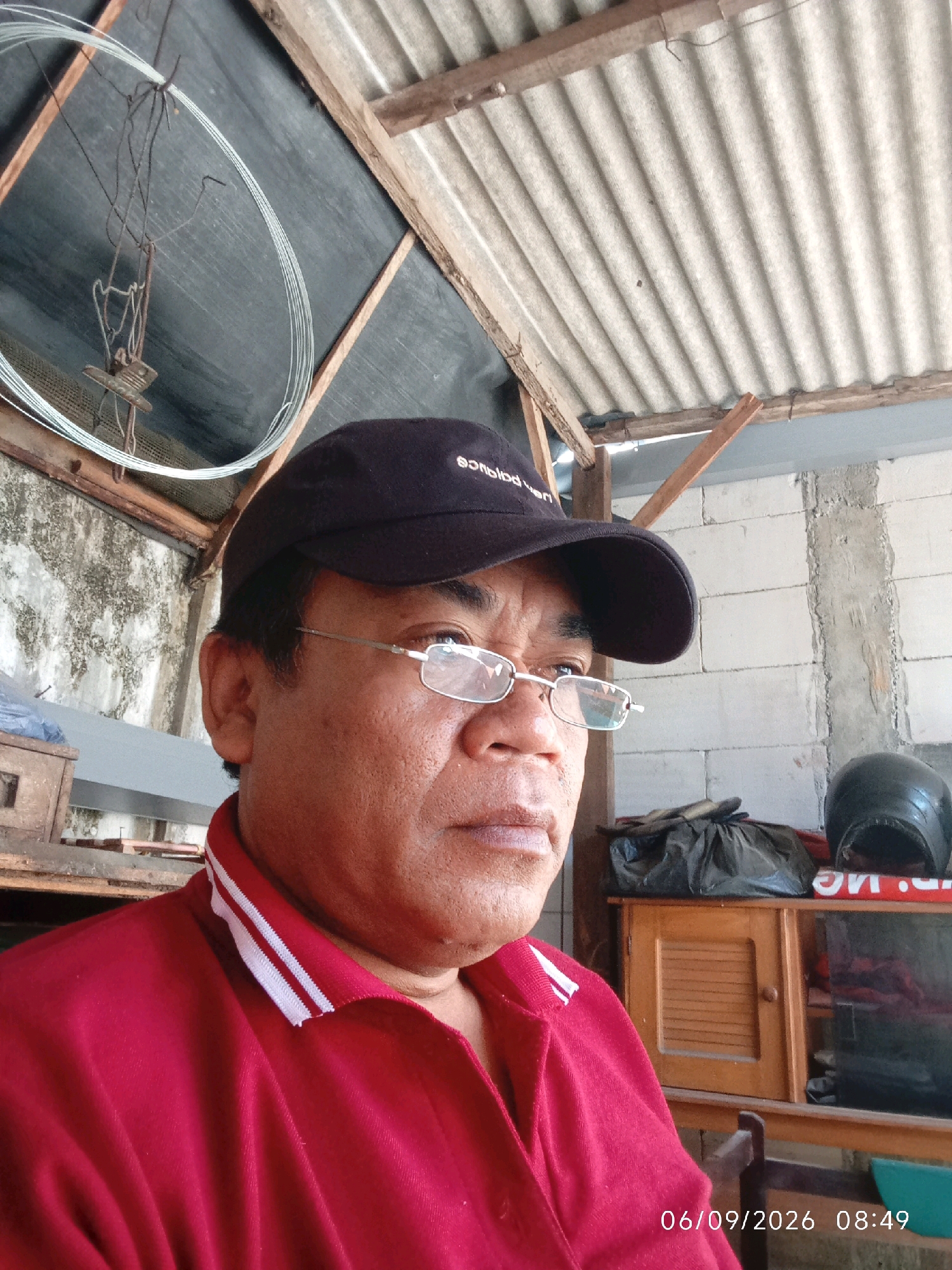 A middle-aged Indonesian man wearing a red polo shirt and a black baseball cap is pictured from the chest up, taking a selfie. He is wearing eyeglasses and looking slightly off-camera. Behind him, the scene appears to be outdoors or in an open-air structure, with a corrugated metal roof supported by wooden beams. The background shows a concrete block wall, a wooden cabinet with louvered doors, and a black motorcycle helmet. The timestamp in the lower right corner indicates the photo was taken on September 6, 2026, at 08:49. The image appears to be taken in Ringinanom, Indonesia, a location characterized by similar rustic, open-air structures and a humid climate conducive to such settings.