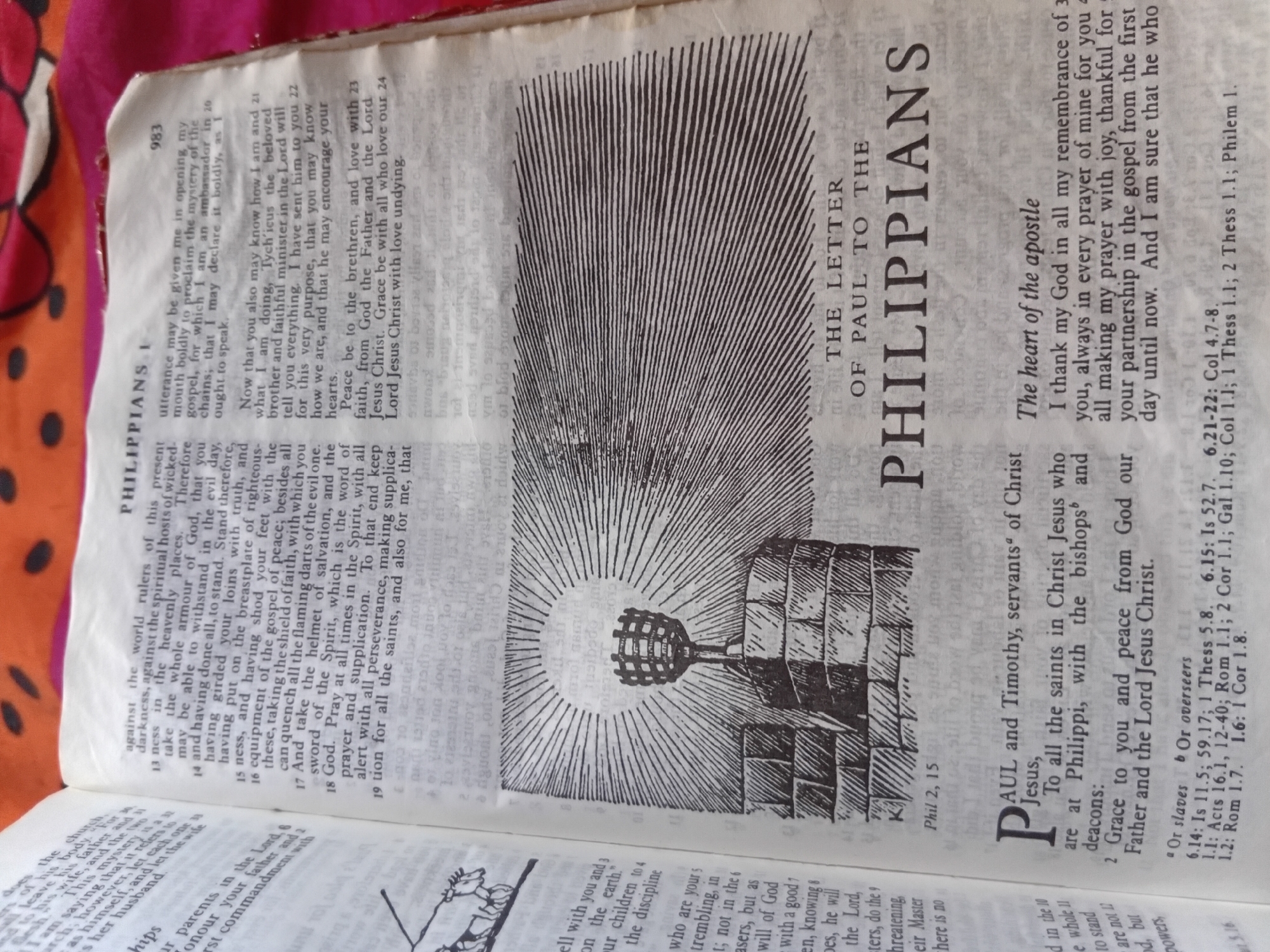 This is a close-up, overhead shot of an open Bible. The page is yellowed with age and the text is printed in black. The Bible is lying on a dark pink fabric with a floral pattern.
The right-hand page contains the text of Philippians 1:3-24, with a large, stylized initial "P" at the beginning of the chapter. The chapter is illustrated with a woodcut of a torch on a stand, emitting rays of light, against a brick wall. The left-hand page contains the text of Ephesians 6:10-24, also with an initial "P" and an illustration of a bull.
Visible text includes:
- "PHILIPPIANS I"
- "THE LETTER OF PAUL TO THE PHILIPPIANS"
- "Phil 2, 15"
- Chapter and verse numbers (e.g., 13, 14, 15, 16, 17, 18, 19, 20, 21, 22, 23, 24)
- "983" (likely a page number)
- Footnotes and cross-references (e.g., "Or slaves", "Or overseers", "Is 11.5", "1 Thess 5.8")
- "The heart of the apostle"
- "P" (large initial letter)
- "AUL and Timothy, servants of Christ Jesus,"
- "To all the saints in Christ Jesus who are at Philippi, with the bishops and deacons:"
- "Grace to you and peace from God our Father and the Lord Jesus Christ."
The lighting is slightly uneven, suggesting it might be indoor lighting or natural light from a window. The overall impression is one of a well-used, older book.