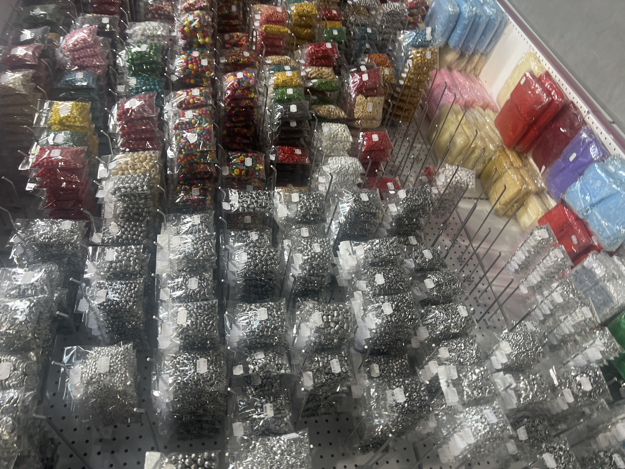 The image shows a well-stocked display of beads and other small craft items, predominantly in clear plastic packaging, hanging from metal rods on a pegboard or similar display system. The merchandise includes beads of various colors (red, green, yellow, blue, pink, white, silver, and multicolored), and also what appear to be small metal fasteners, organized by color and type. The items are presumably for sale. The location is most likely a retail store specializing in crafts and DIY supplies. It could potentially be located in Burgas, Bulgaria based on your request. The environment is well-lit, suggesting an indoor setting. There are no people visible in the image. The focus is purely on the arrangement of the merchandise.