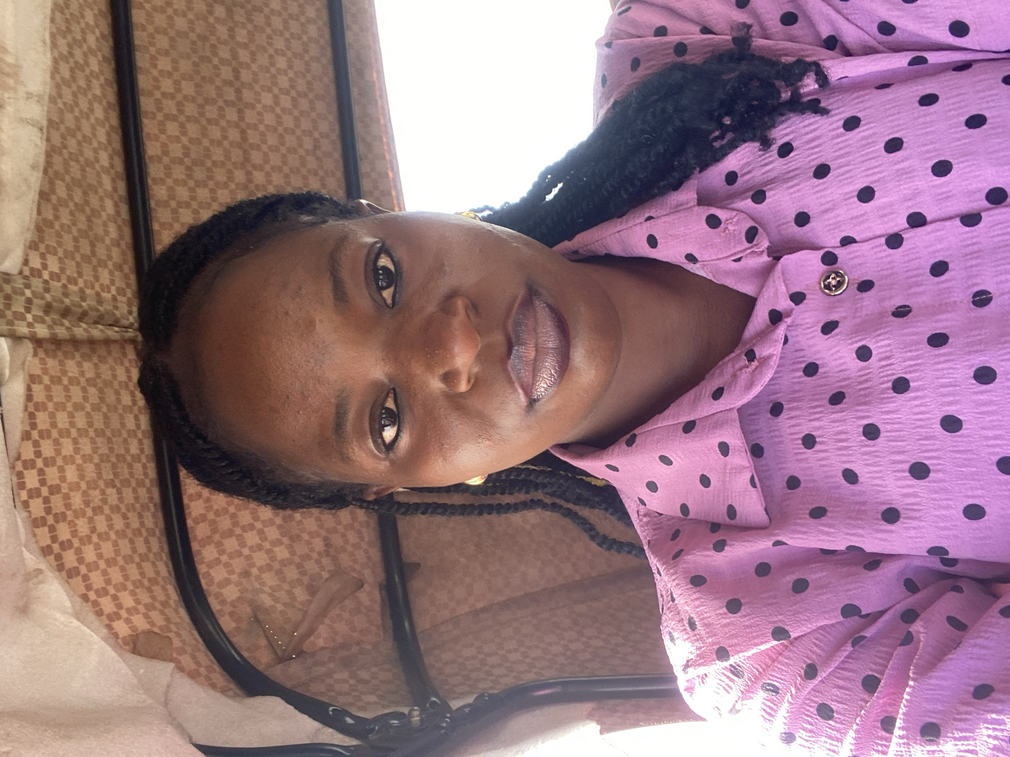 The image shows a close-up, selfie-style shot of a woman inside a vehicle, possibly a tuk-tuk, with a patterned canopy overhead. The woman is of African descent and has dark skin. Her hair is styled in braids that fall over her shoulders. She is wearing a pink button-down shirt with a black polka dot pattern. She has on gold hoop earrings and appears to be wearing dark lipstick. The background shows the interior of the vehicle, including a black metal frame and the textured, beige and brown patterned canopy. The lighting suggests it is daytime and the image has been taken outdoors. There is no indication of the specific location within the city of Du, Nigeria.