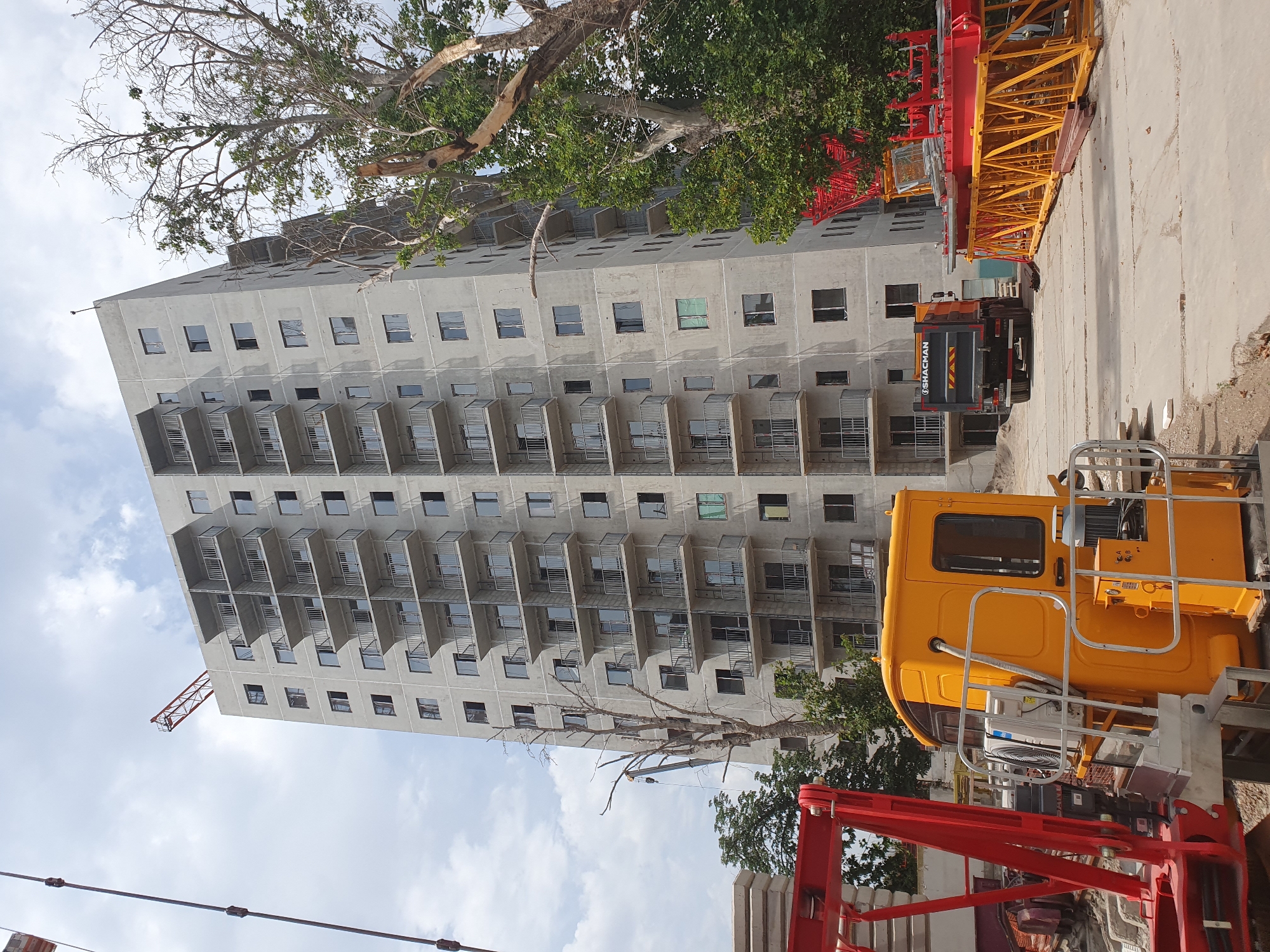 This is an exterior, eye-level, daytime, medium shot of a high-rise apartment building under construction in Tashkent, Uzbekistan. The unfinished concrete building dominates the frame, featuring multiple balconies with metal railings and dark-framed windows. A red crane arm extends from the top left of the building. In the foreground, a yellow construction vehicle cabin with various mechanical components is visible, with red crane components stacked to its right. A dark-colored dump truck with "SHACMAN" branding is parked on the dirt ground to the left of the building. Several bare, dead trees are scattered throughout the scene, contrasting with the green foliage of a large tree on the right side of the image. The sky is partly cloudy.