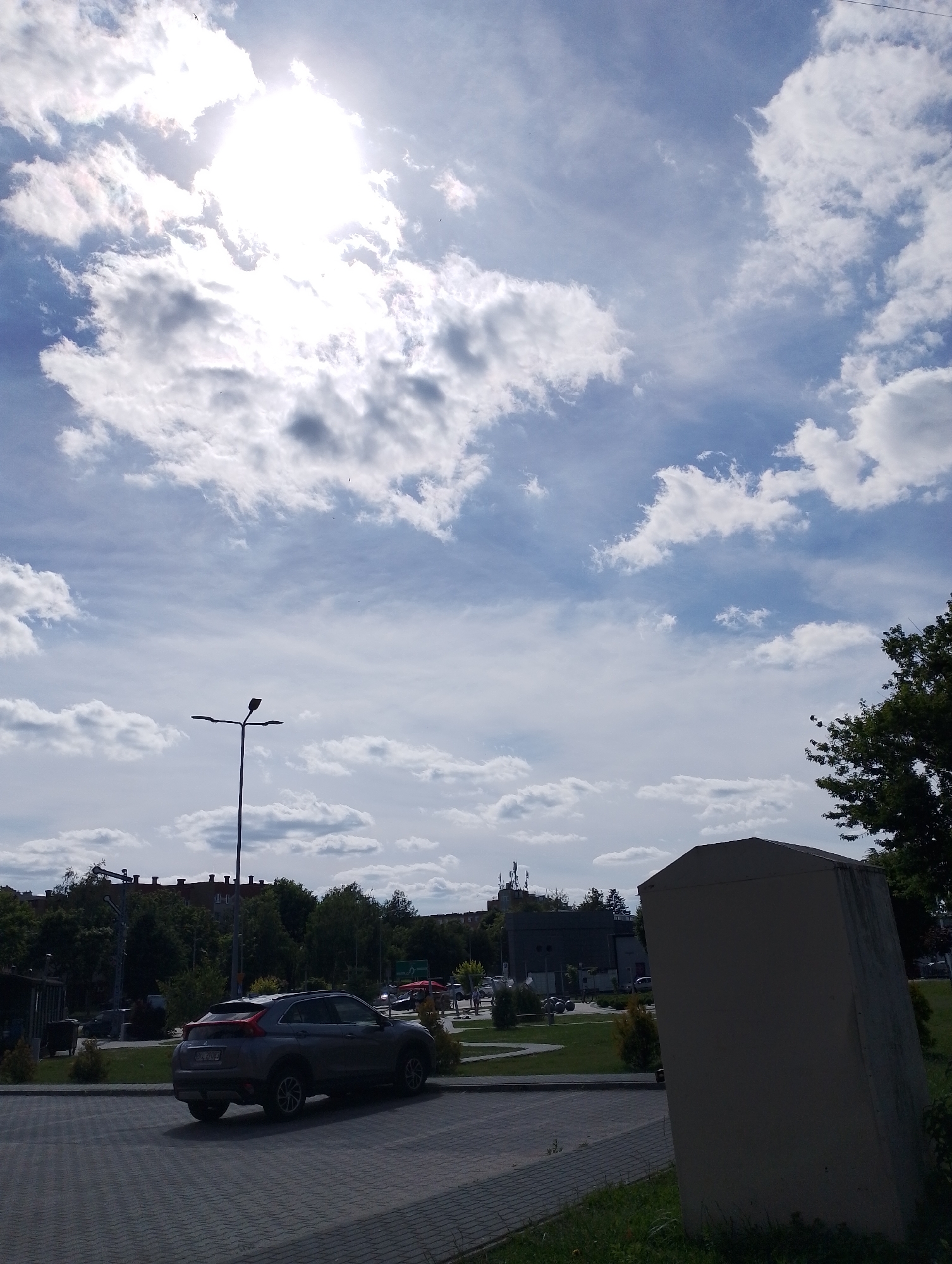 The image shows a street scene under a cloudy sky with the sun partially obscured by clouds. In the foreground, a grey car is parked on a paved area. To the right, a large, light-colored rectangular structure stands on a grassy area. In the background, there are trees, buildings, and a streetlamp. Several other vehicles and some pedestrians are visible in the distance. The overall environment appears to be an urban or suburban setting.