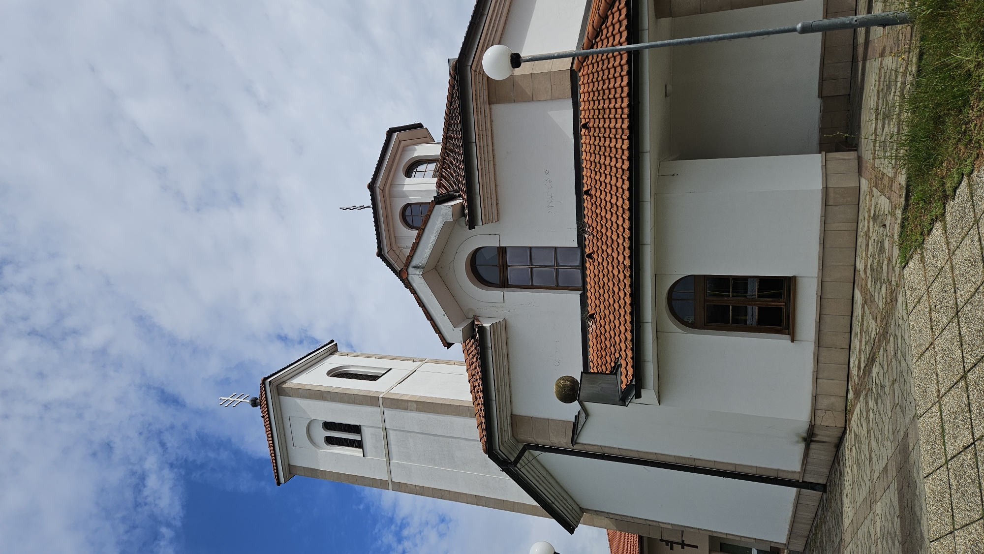 The image shows a white church with a red tiled roof, set against a blue sky with scattered clouds. A tall, white bell tower with a cross on top stands to the left of the main building. The church features arched windows and a circular dome with smaller arched windows. The exterior walls are adorned with light brown stone accents. The foreground shows a paved area with interlocking concrete pavers, and a lamppost with a white globe is visible on the right. The scene appears to be an exterior view of the Church of the Holy Spirit in Prešov, Slovakia, a prominent religious landmark in the city.